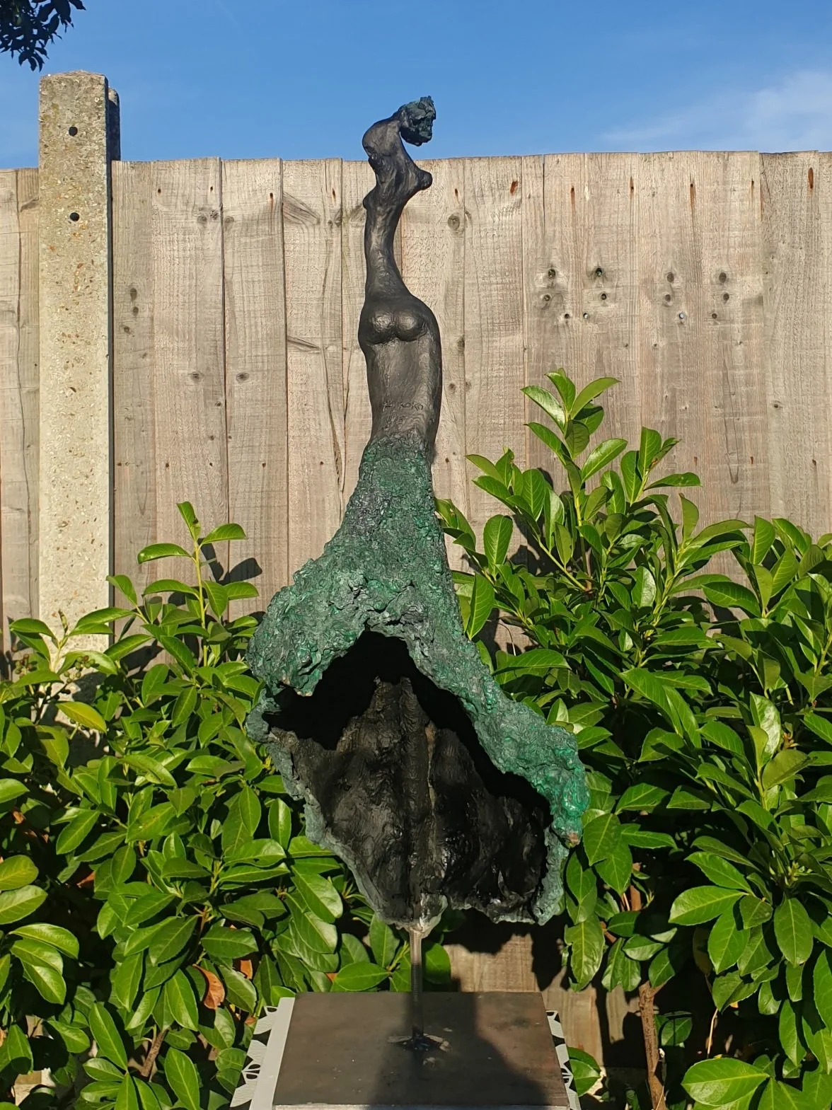 Sculpture of a tall, slender woman with a long neck and minimal facial features, mounted on a stand outdoors, against a wooden fence with green bushes and a clear blue sky in the background.