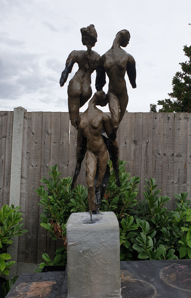 A bronze sculpture of three intertwined nude female figures standing on a rectangular stone base, with a wooden fence and green shrubs in the background.