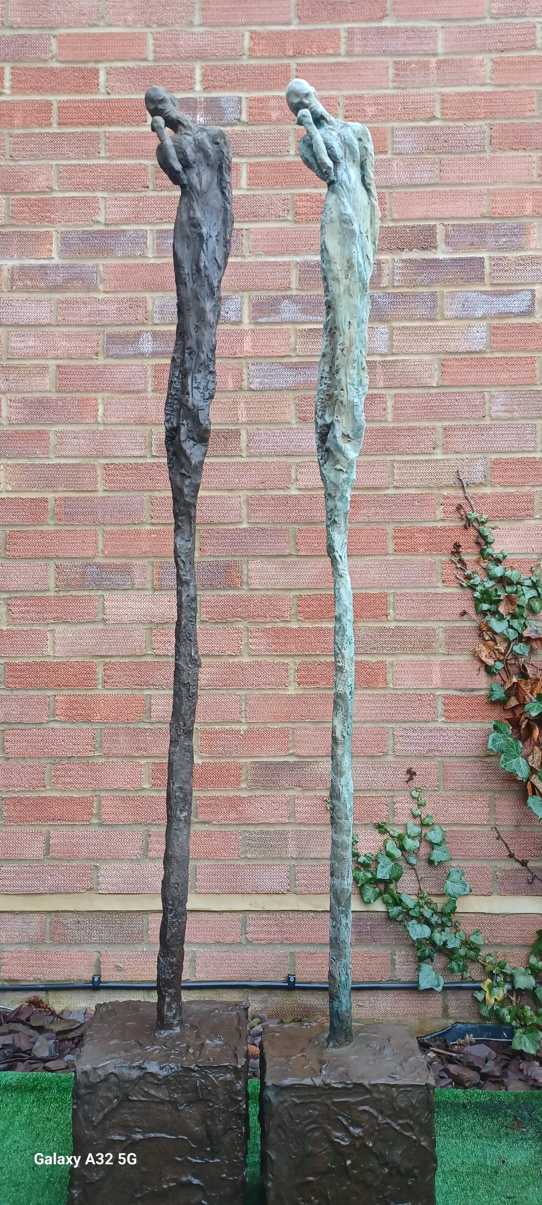 Tall, abstract bronze sculptures of elongated human figures with one hand on their faces are placed against a brick wall. The sculptures are mounted on square bases, with some greenery on the right side.