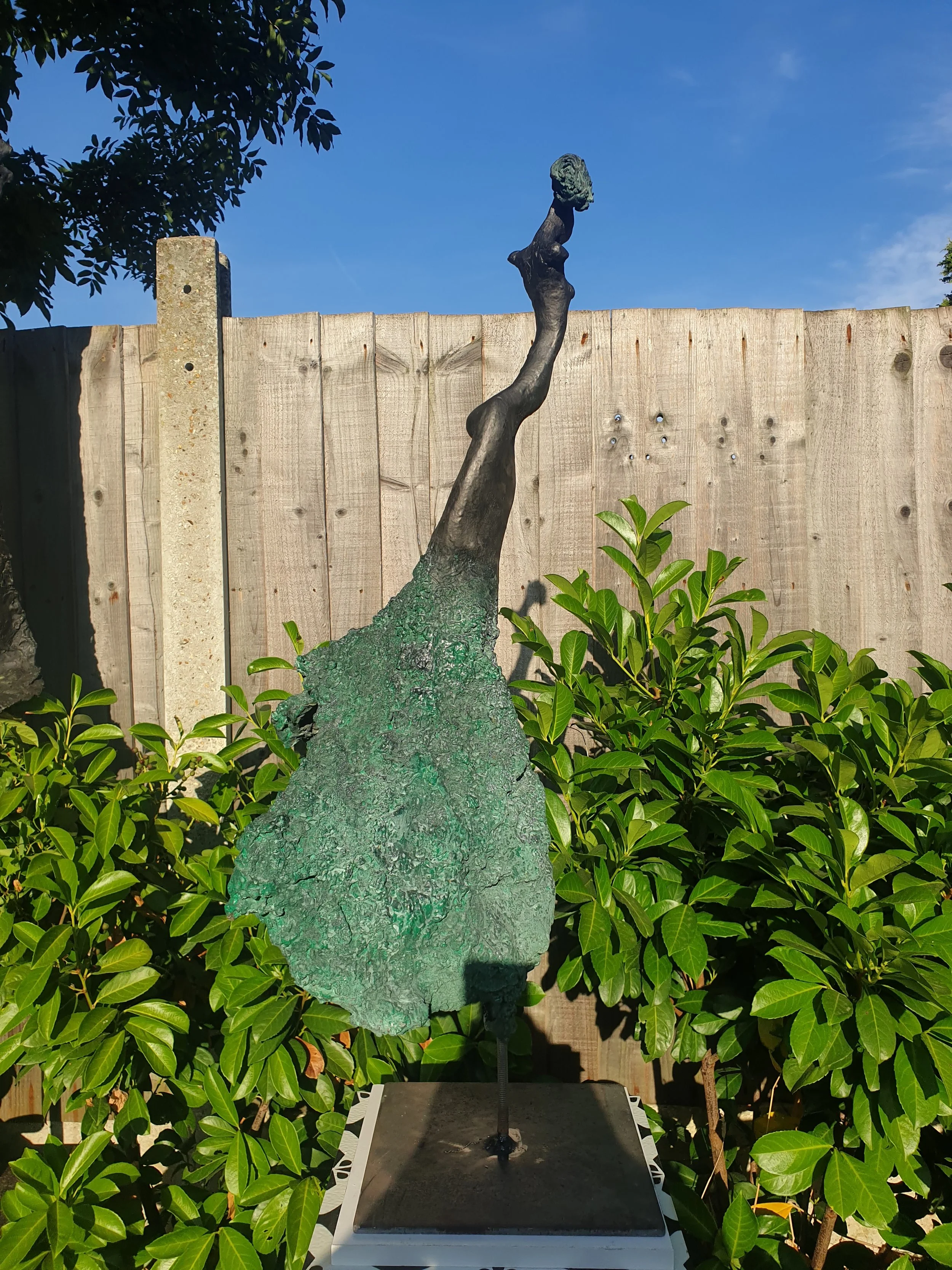 Sculpture of a stylized heron or crane with a long neck, made of metal with a patina, standing on a base outdoors surrounded by green bushes and a wooden fence, with a clear blue sky in the background.