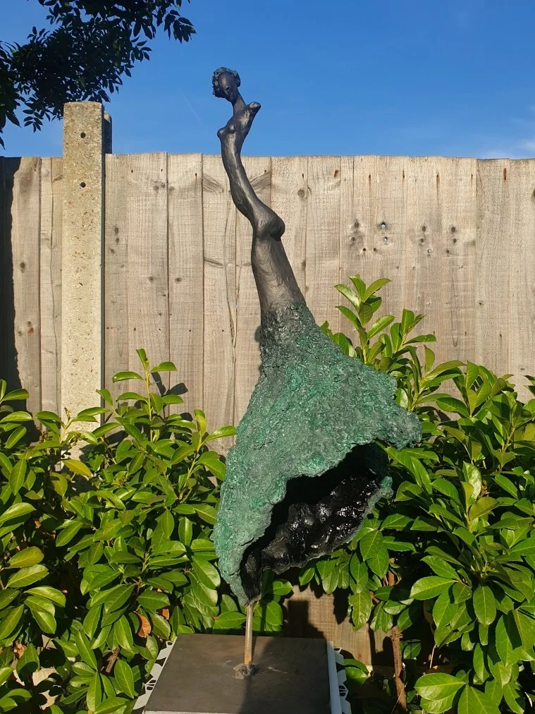 A sculpture of a tall, slender female figure with elongated limbs and neck, situated outdoors against a wooden fence and green bushes, under a clear blue sky.