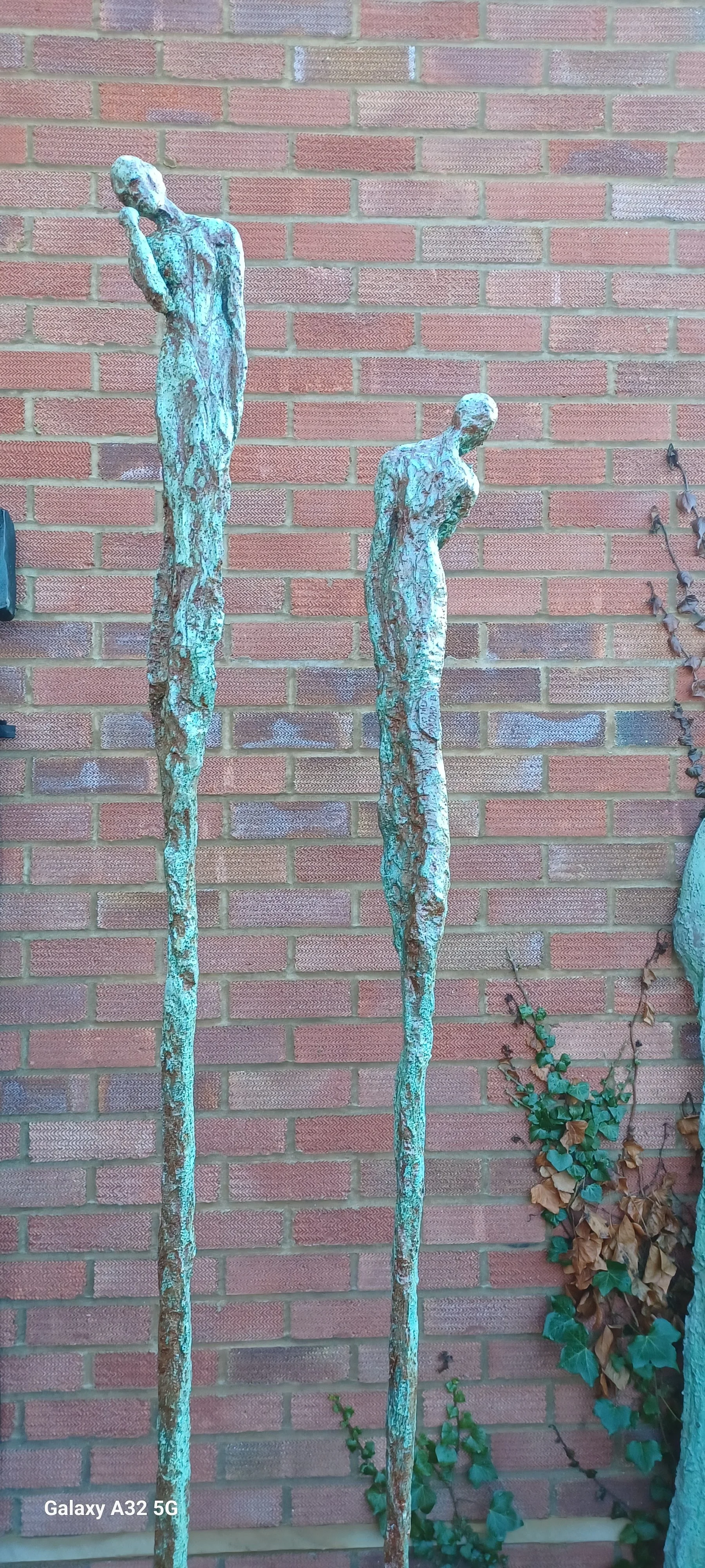 Two tall, weathered bronze sculptures of slender, elongated human figures standing against a brick wall, with one figure bowing and the other holding a hand to its face, surrounded by some creeping vines.