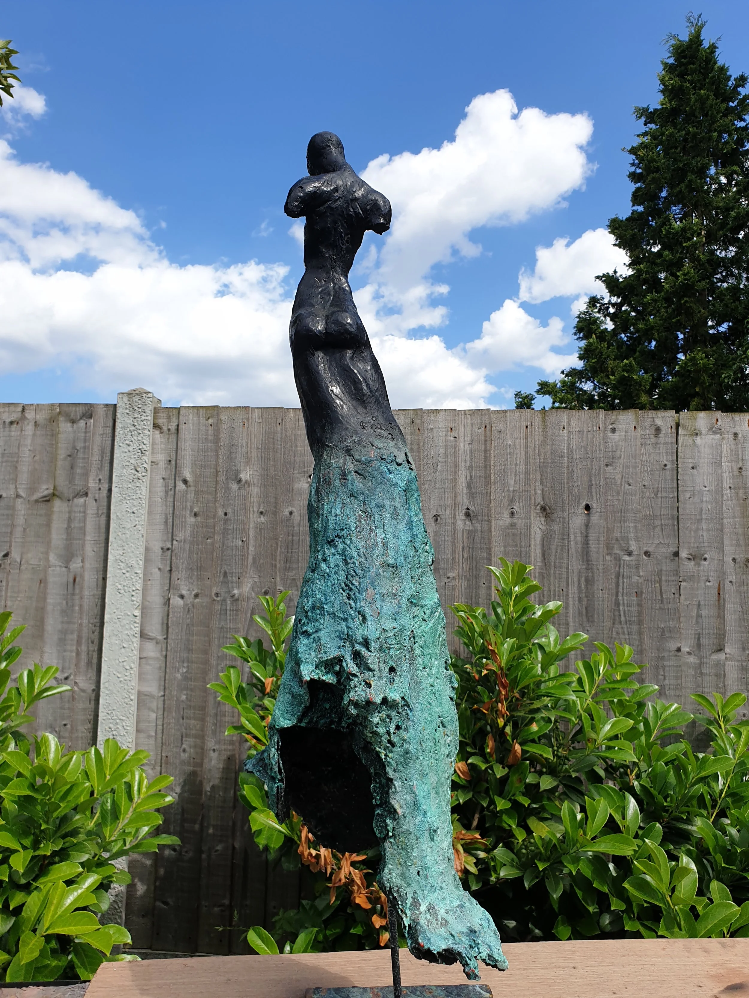 A sculpture of a woman with arms crossed, made of dark and greenish material, standing outdoors in front of a wooden fence, green bushes, and a blue sky with white clouds.