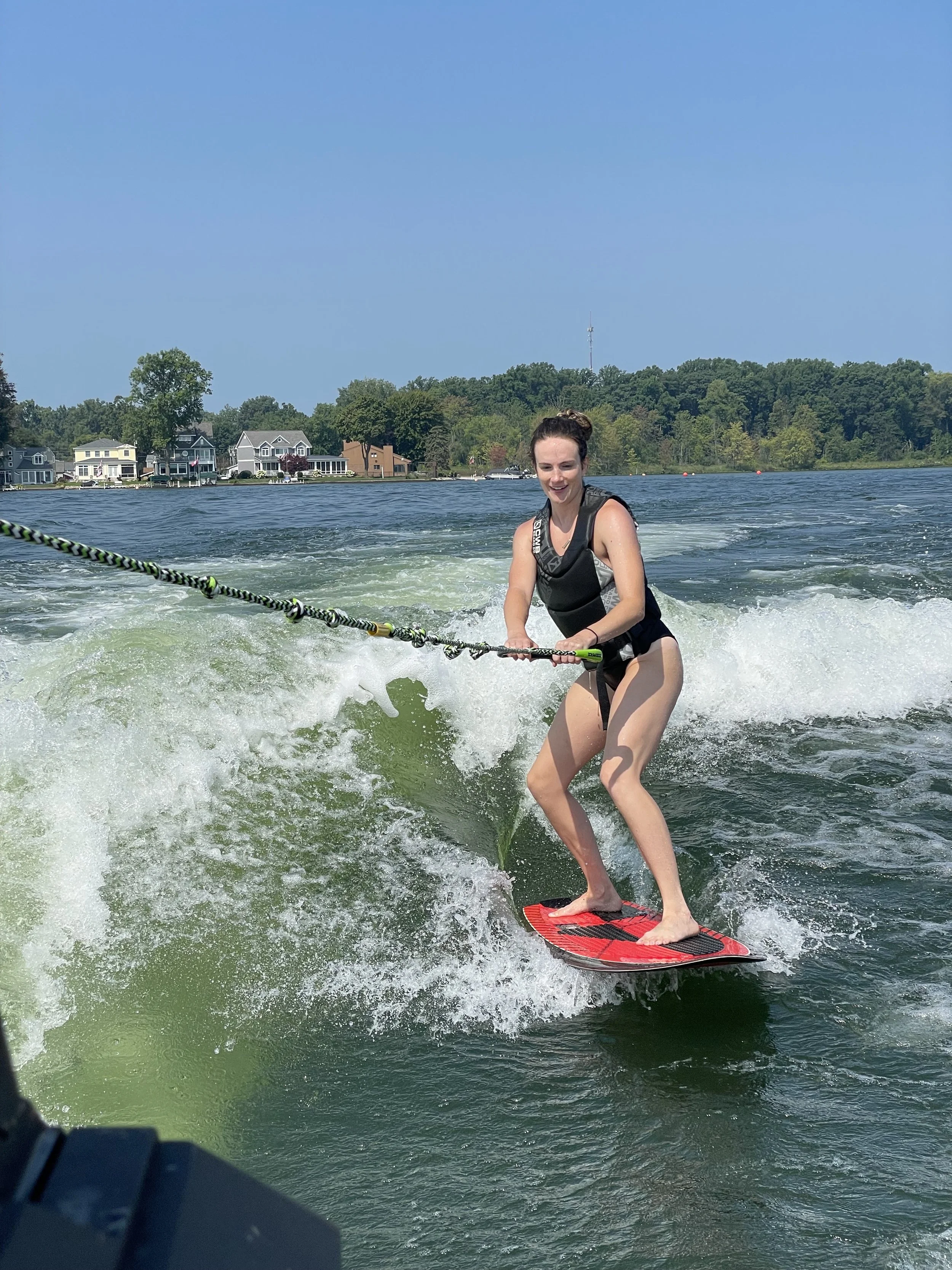 A young woman wearing a black life jacket and black swimsuit is wakeboarding on a lake. She is holding onto a tow rope and standing on a red wakeboard, smiling as she rides a wave with houses and trees in the background under a clear blue sky.