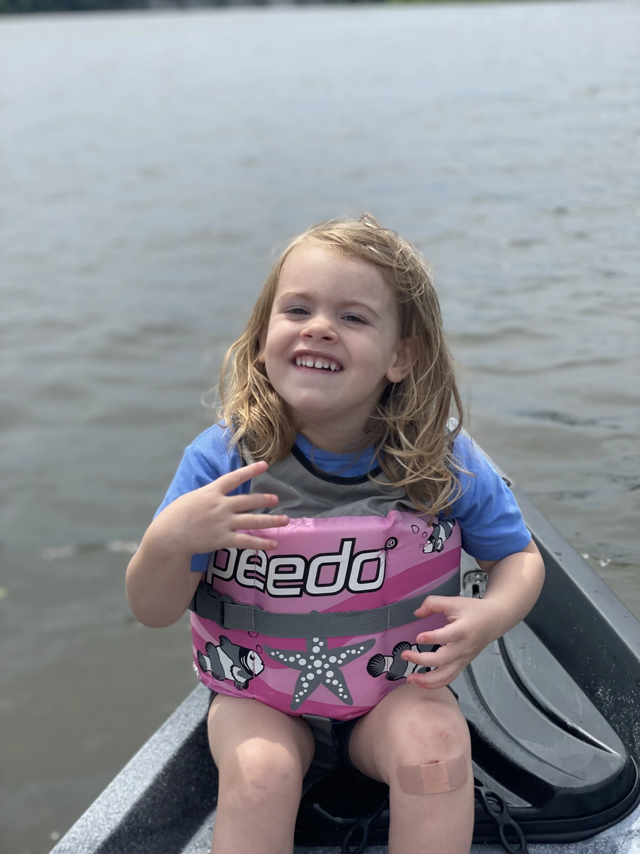 A smiling young girl with curly hair wearing a pink life jacket and blue shirt, sitting in a boat on a body of water.