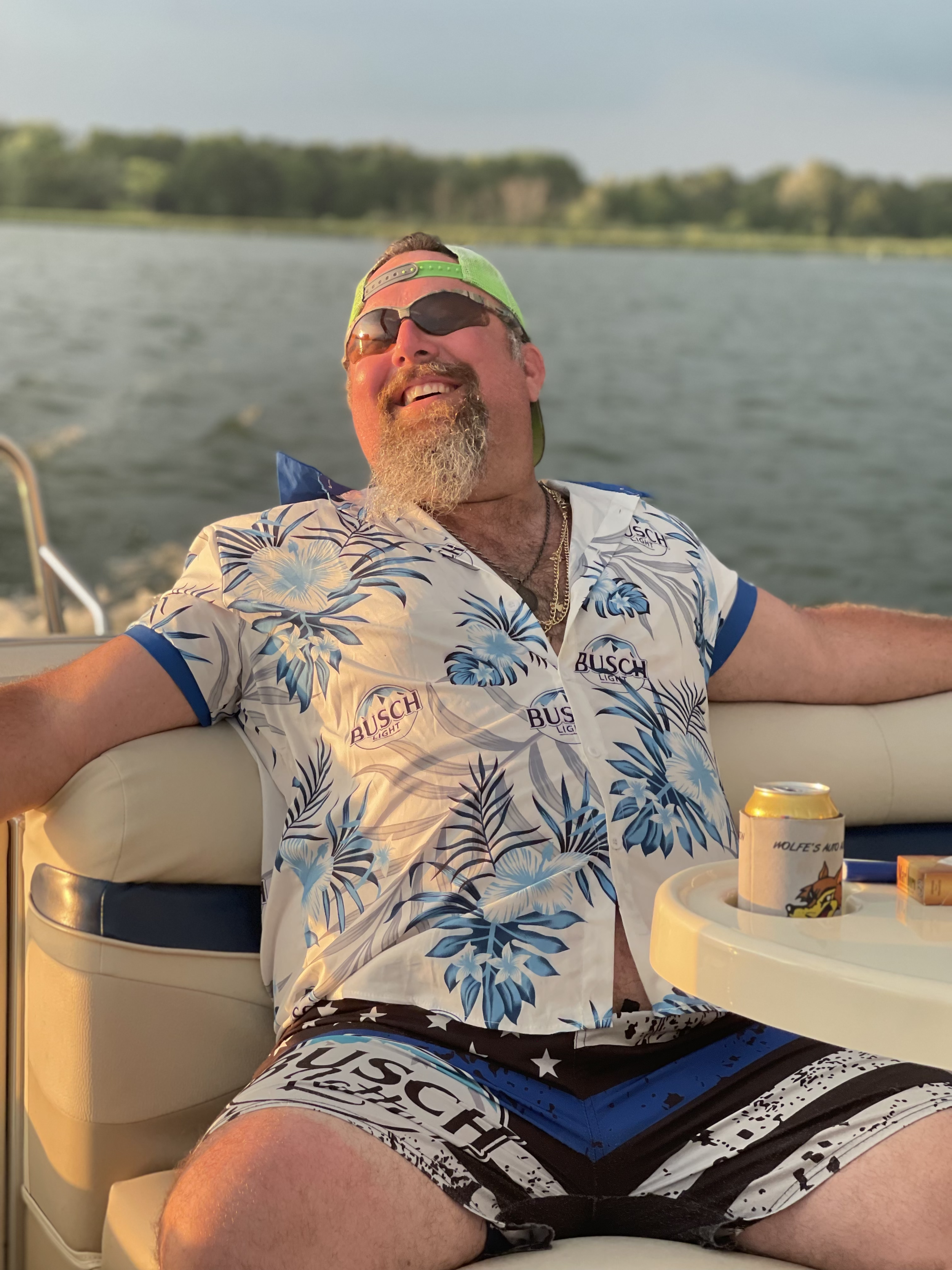 A man relaxing on a boat wearing a floral Hawaiian shirt, patterned shorts, sunglasses, and a backward cap, smiling with water and trees in the background.
