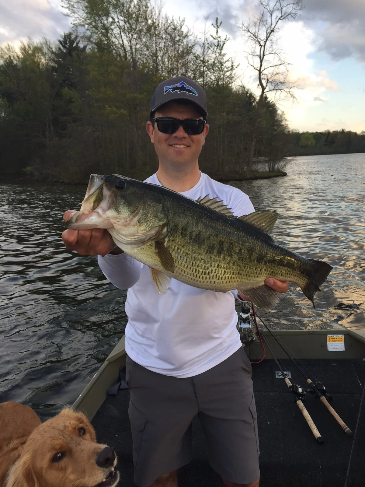 Man wearing sunglasses and a cap, holding a large bass fish on a boat on a lake with trees in the background and a dog in the foreground.
