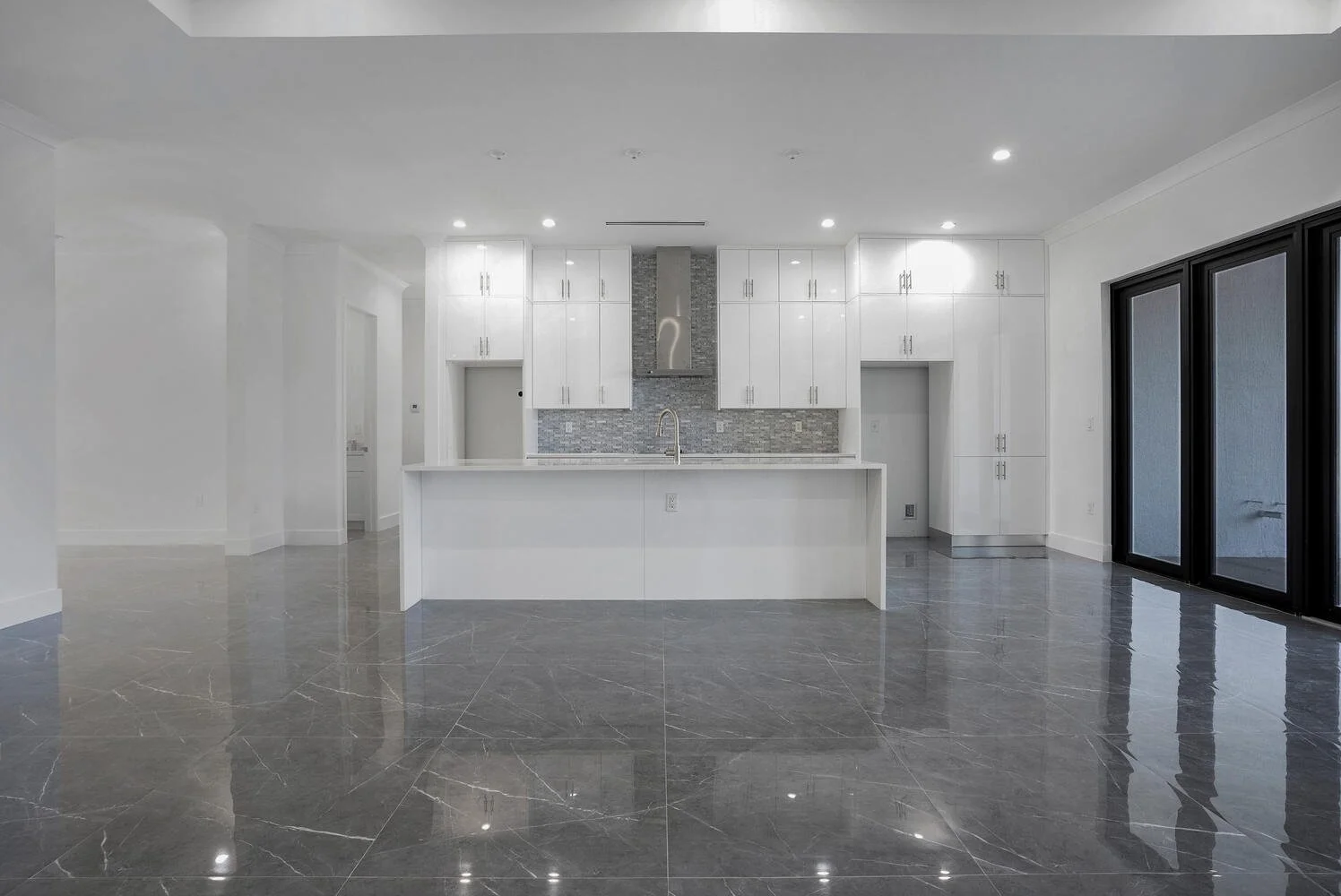 Modern, empty white kitchen with marble floors, island with sink, white cabinets, gray backsplash, and sliding glass doors.