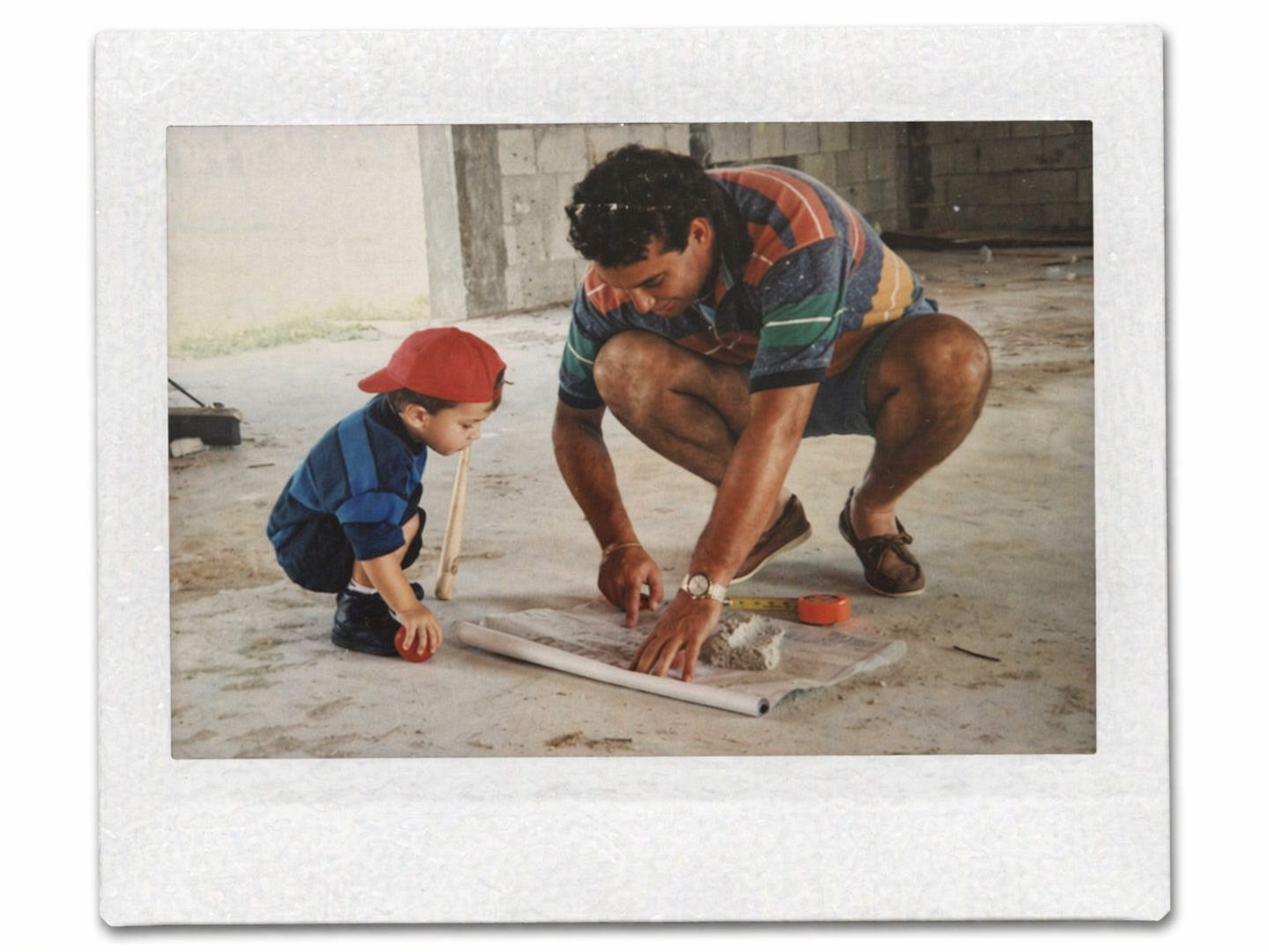 A man and a young boy crouch on the floor, working on a project with a blueprint or plan, in what appears to be an unfinished or under construction building.