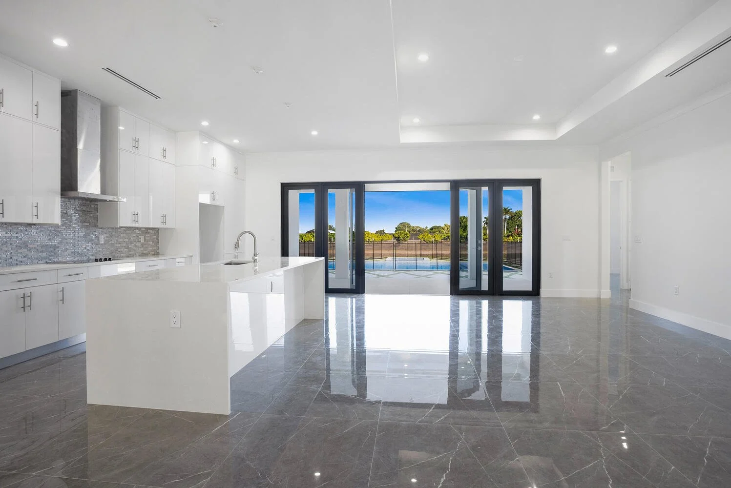 Empty modern kitchen with white cabinets, marble island, gray tiled backsplash, and large glass sliding doors leading to a backyard with a pool and trees, under a blue sky.
