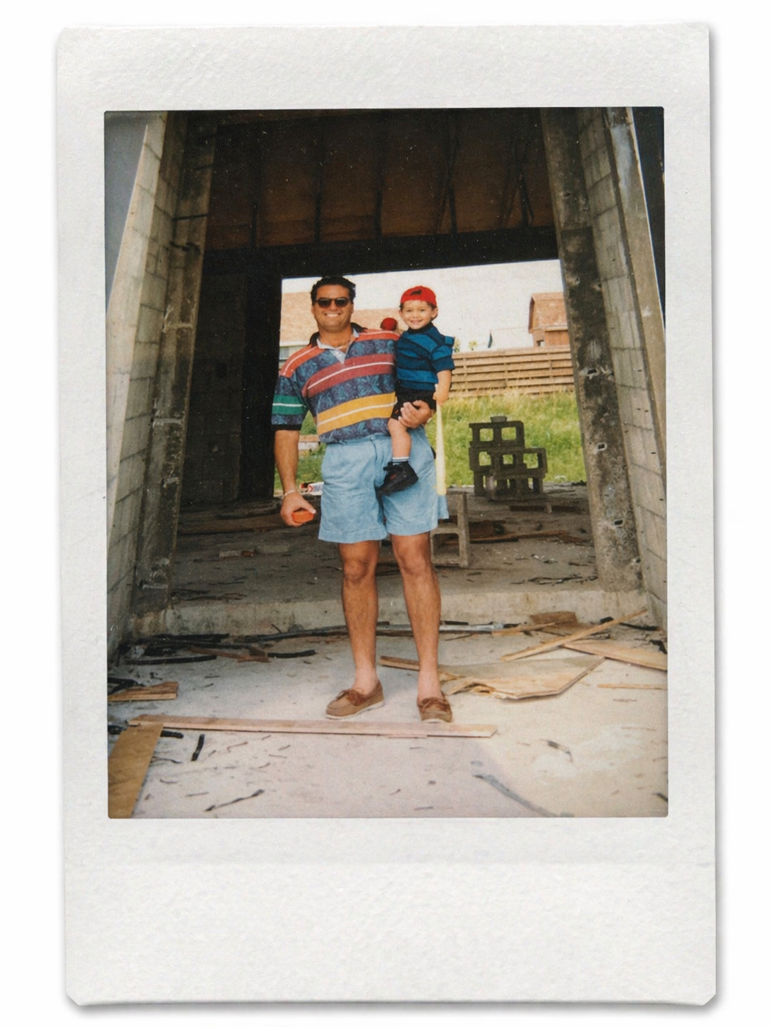 A man in colorful striped shirt and shorts holding a young boy in a red cap and striped shirt inside a partially built wooden structure.