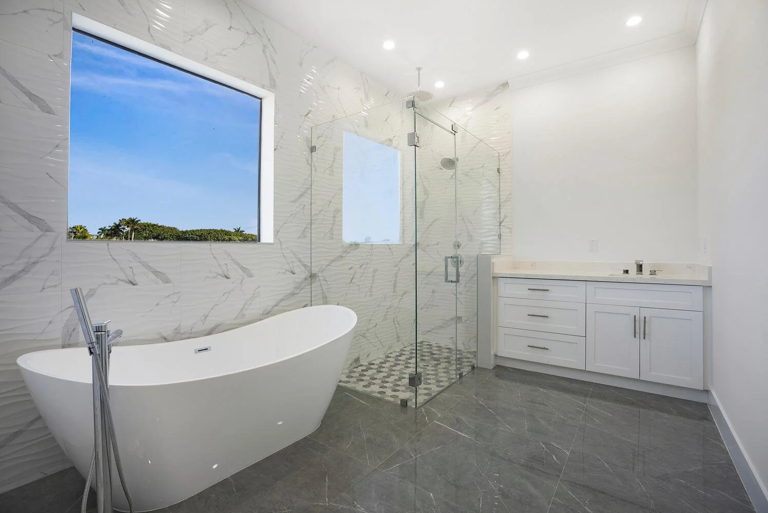 Modern bathroom featuring a white freestanding bathtub, a glass-enclosed shower area with marble tiles, a white vanity with storage, a large window showing a clear blue sky, and gray tiled flooring.