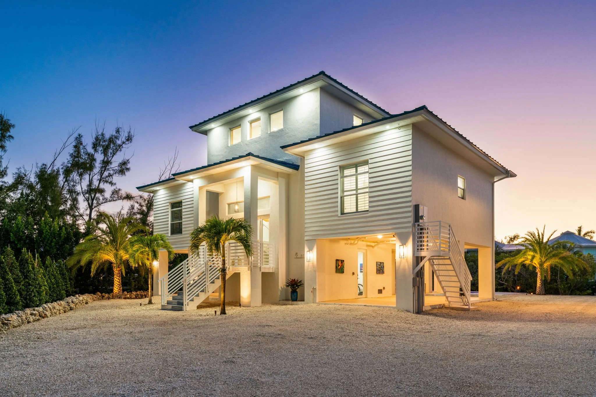 A modern white house in Key Largo with multiple levels and large windows, illuminated at dusk, with palm trees and landscaped yard.