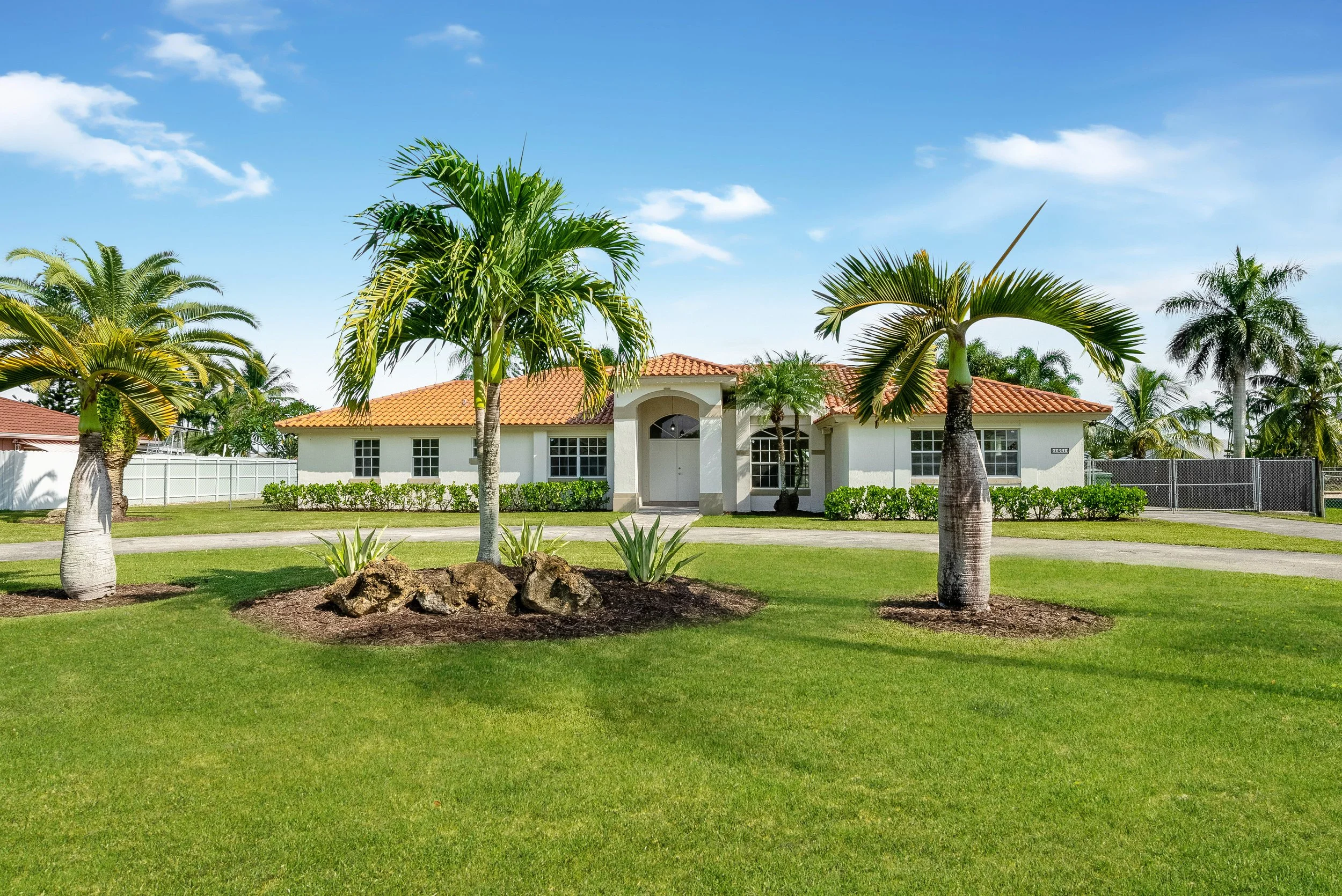 A white house with an orange tiled roof, surrounded by palm trees and green lawn under a blue sky with wispy clouds.