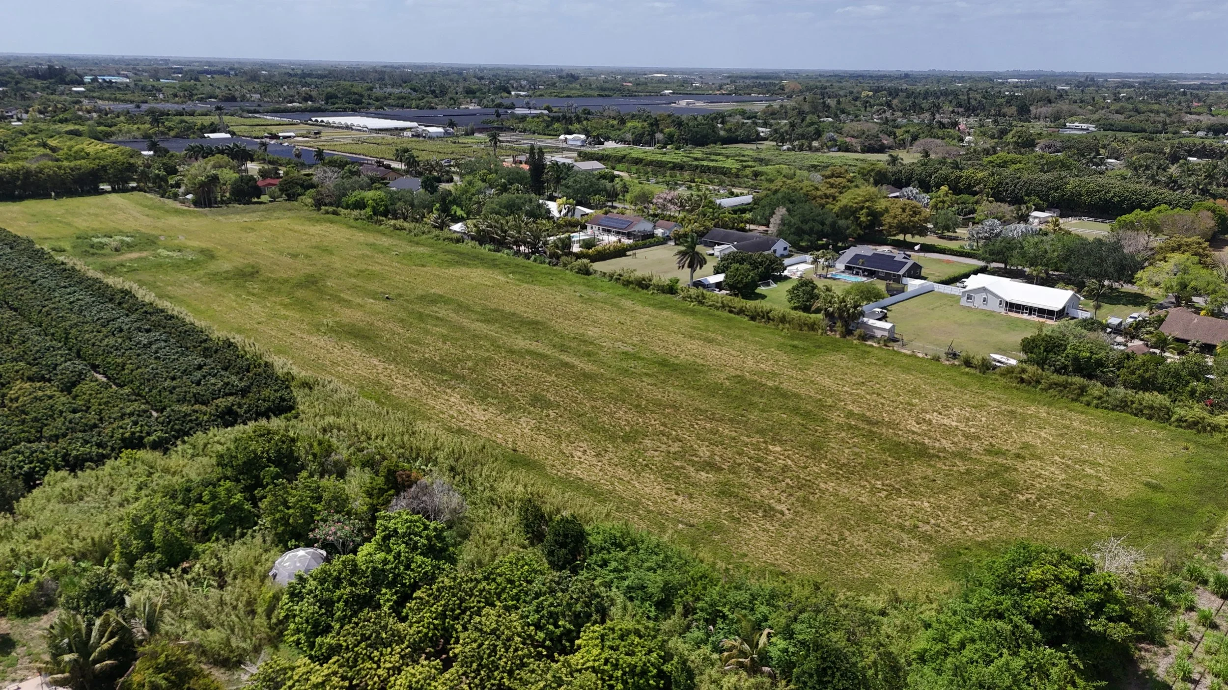 Aerial view of a suburban neighborhood with large green yards, trees, and scattered houses, surrounded by lush greenery and open fields, under a partly cloudy sky.