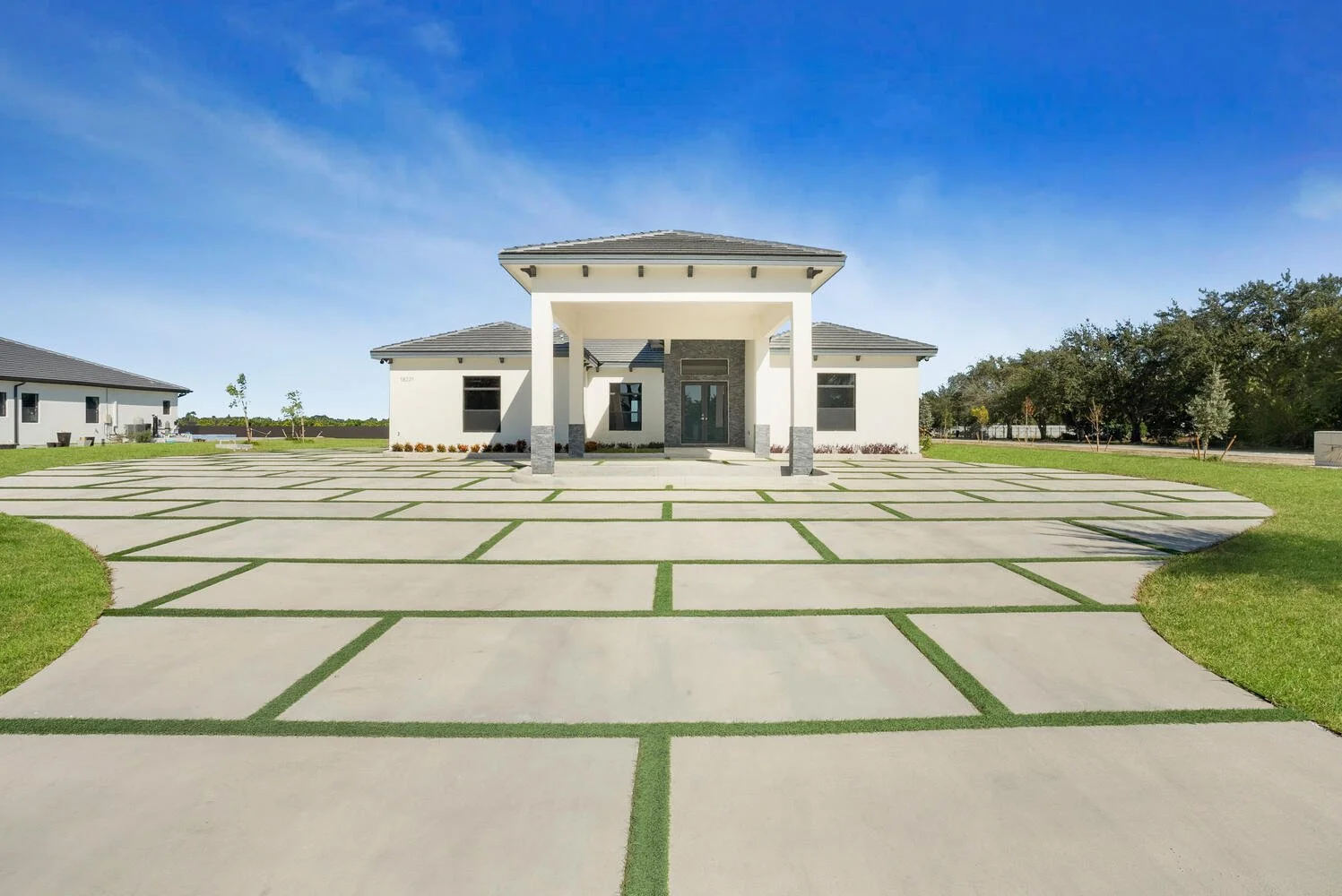 Front view of a modern single-story house with a spacious layout, concrete driveway with grass-lined geometric patterns, and a clear blue sky.