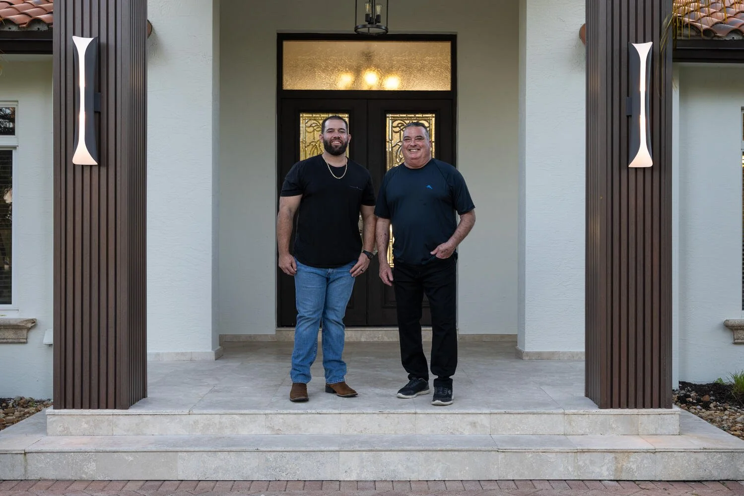 Two men standing on the front steps of their house, smiling, with a white exterior, black front door with decorative glass, and modern outdoor lighting.