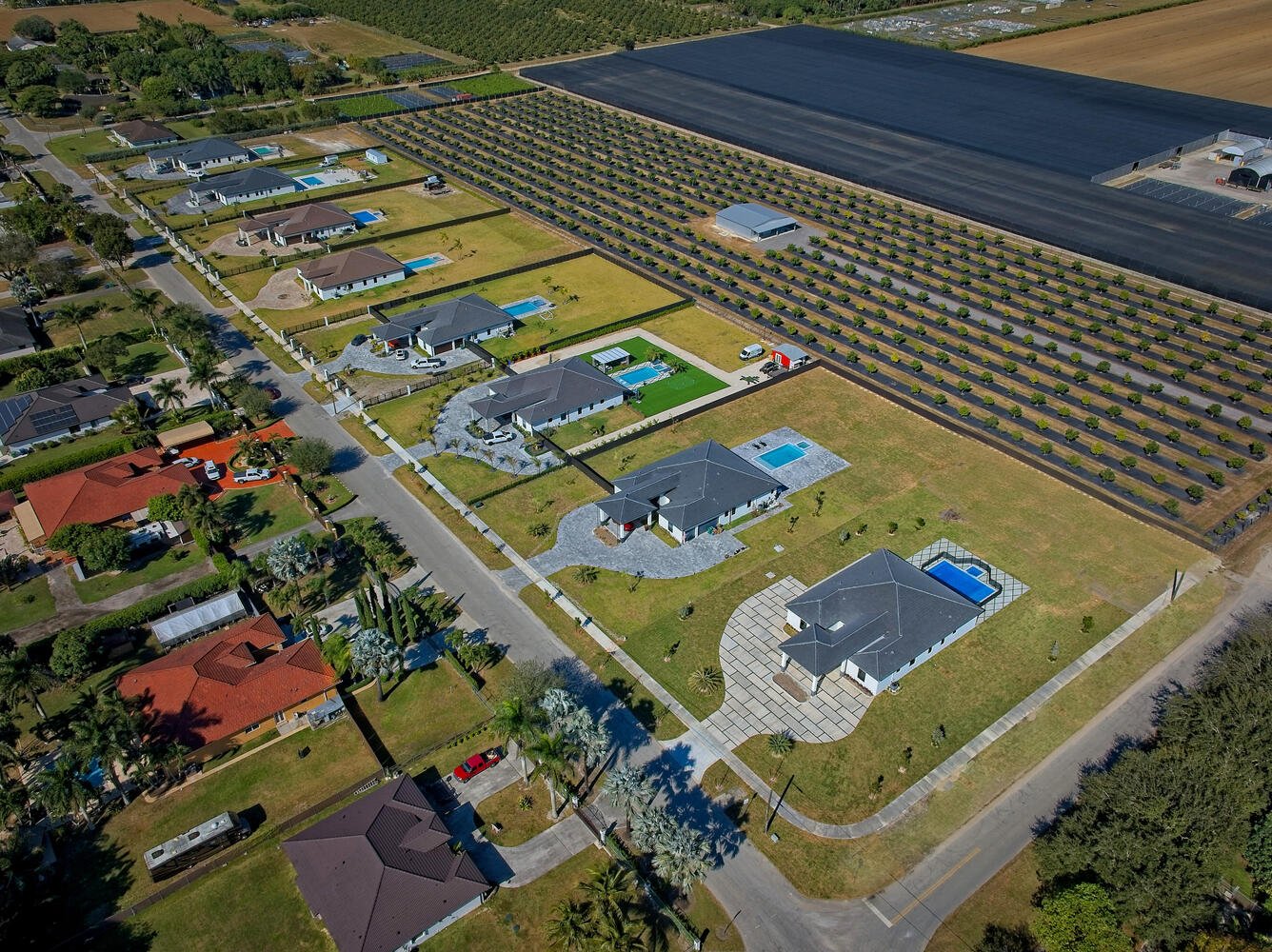 Aerial view of a residential neighborhood with single-family houses, each with a yard and some with swimming pools, alongside a large farm or nursery with rows of plants for cultivation.