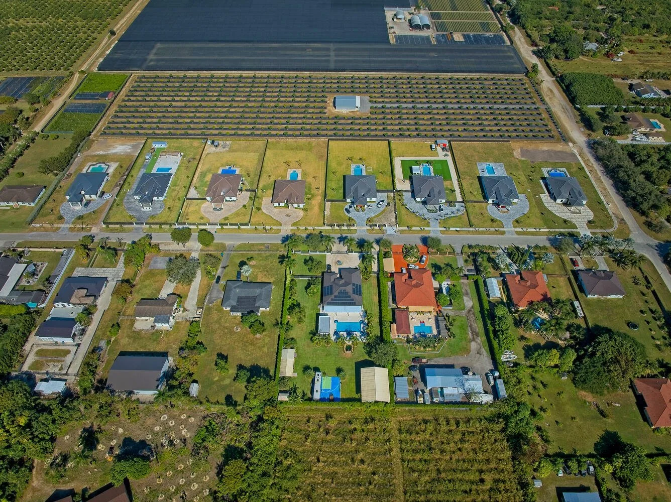 Aerial view of suburban neighborhood with houses, swimming pools, and manicured lawns, surrounded by farmland and green fields.