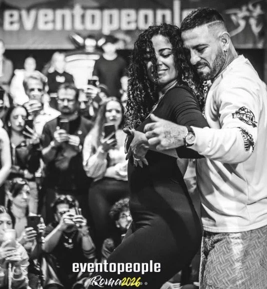La Alemana and Daniel dance closely together in front of an audience holding up phones. The background shows a large sign that reads 'eventpeople'.