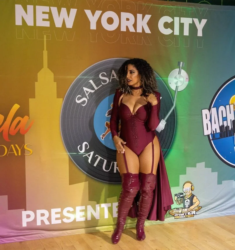 La Alemana with curly hair wearing a burgundy bodysuit, thigh-high boots, and a long cape, standing in front of a colorful backdrop with the text 'New York City' and various event logos.