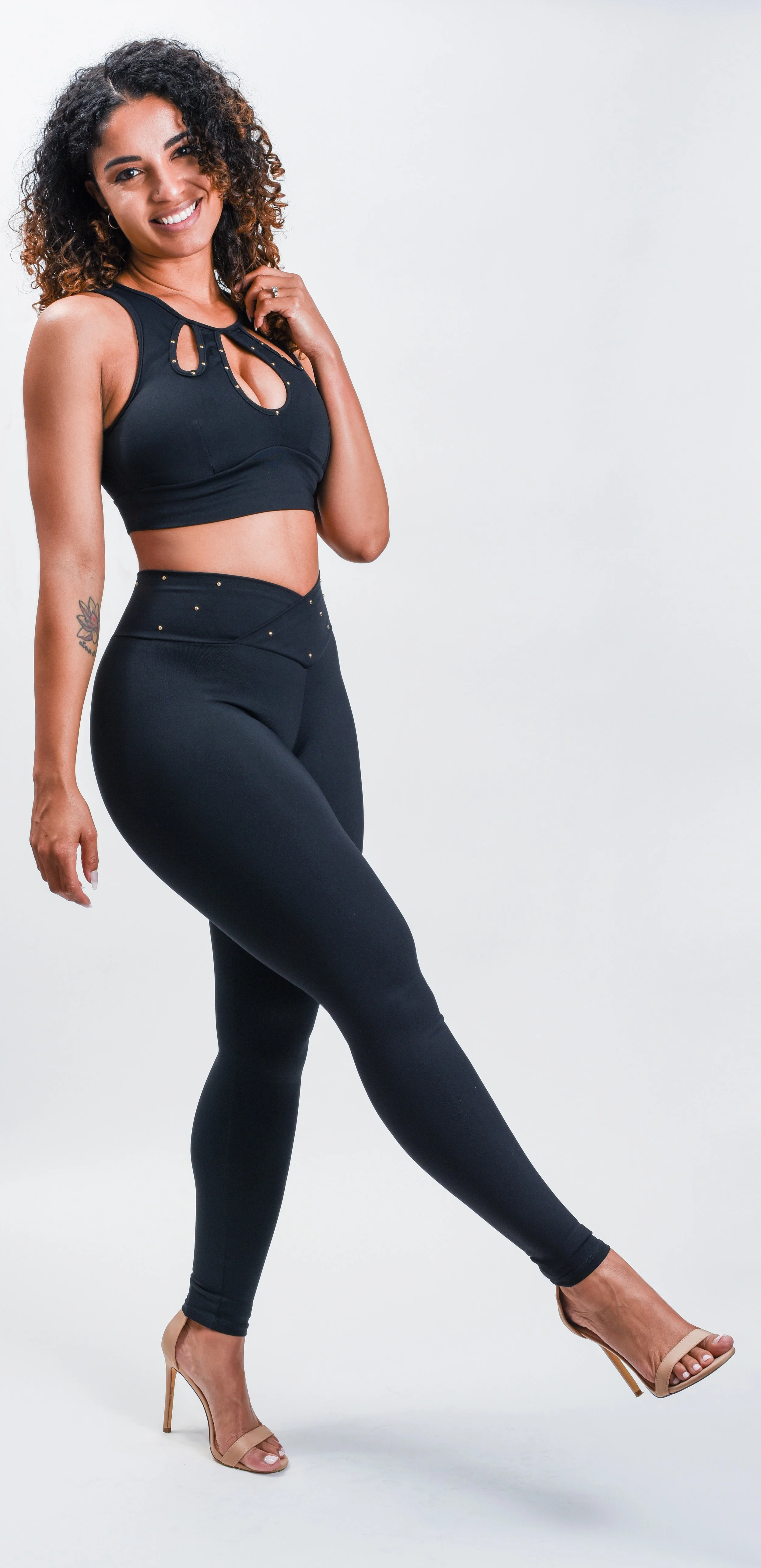 La Alemana with curly hair wearing a black sports bra and matching black leggings, beige high heels, and smiling while posing against a plain white background.