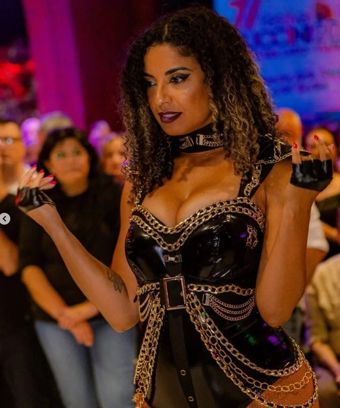 La Alemana dancing with curly hair dressed in a black leather outfit with chain details, standing at an event with a blurred audience in the background.