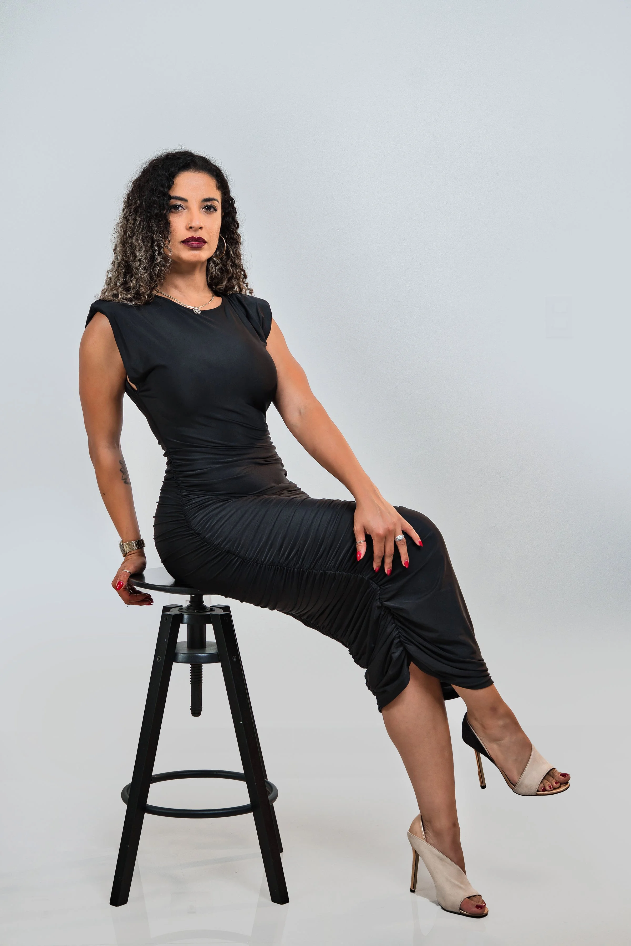 La Alemana with curly dark hair and dark lipstick sits on a black stool against a plain light gray background. She wears a black midi dress with ruched detailing and beige high heels with metallic heels.