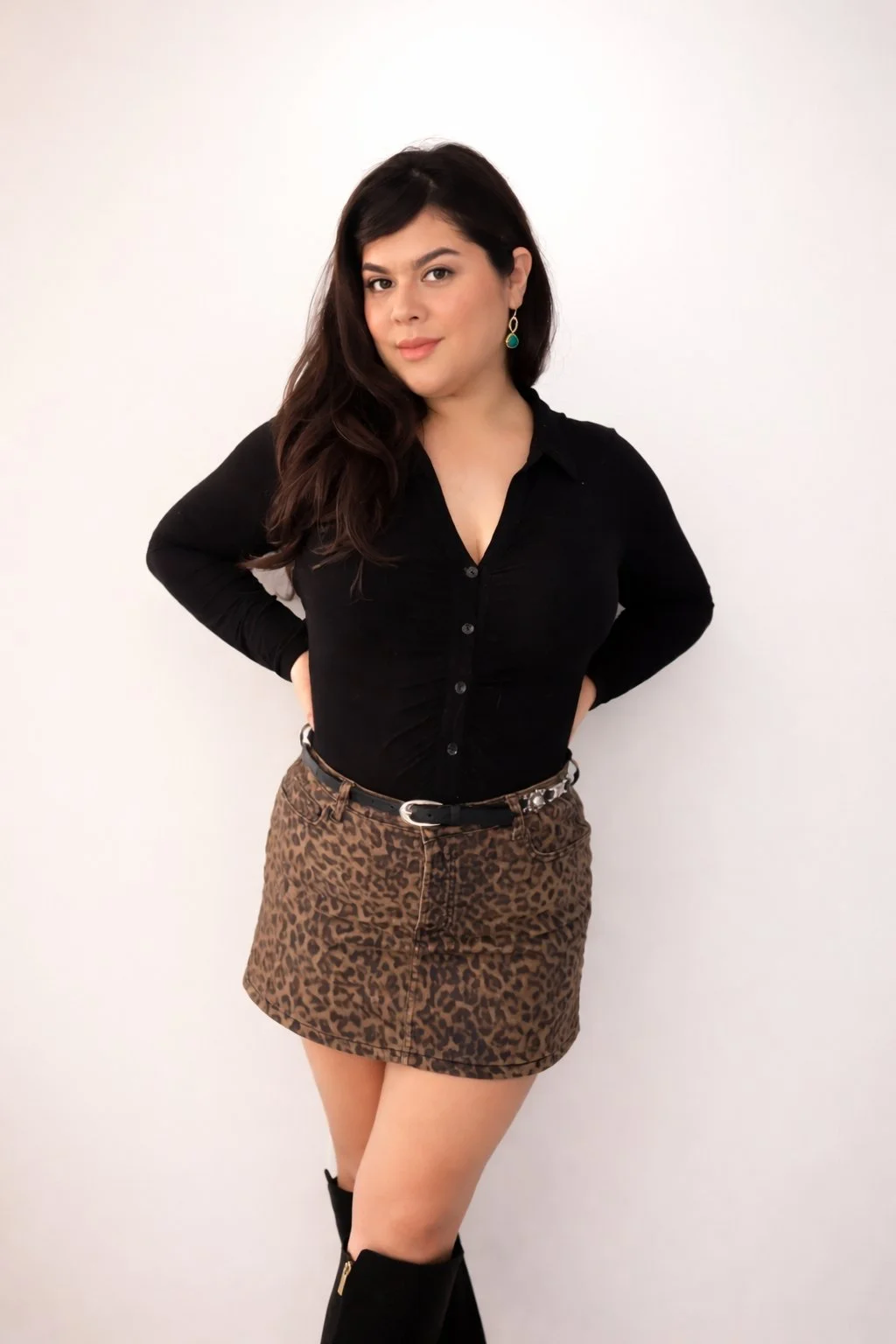 A young woman with long dark hair, wearing a black button-up top, a leopard print skirt, and knee-high black boots, standing against a plain white background.