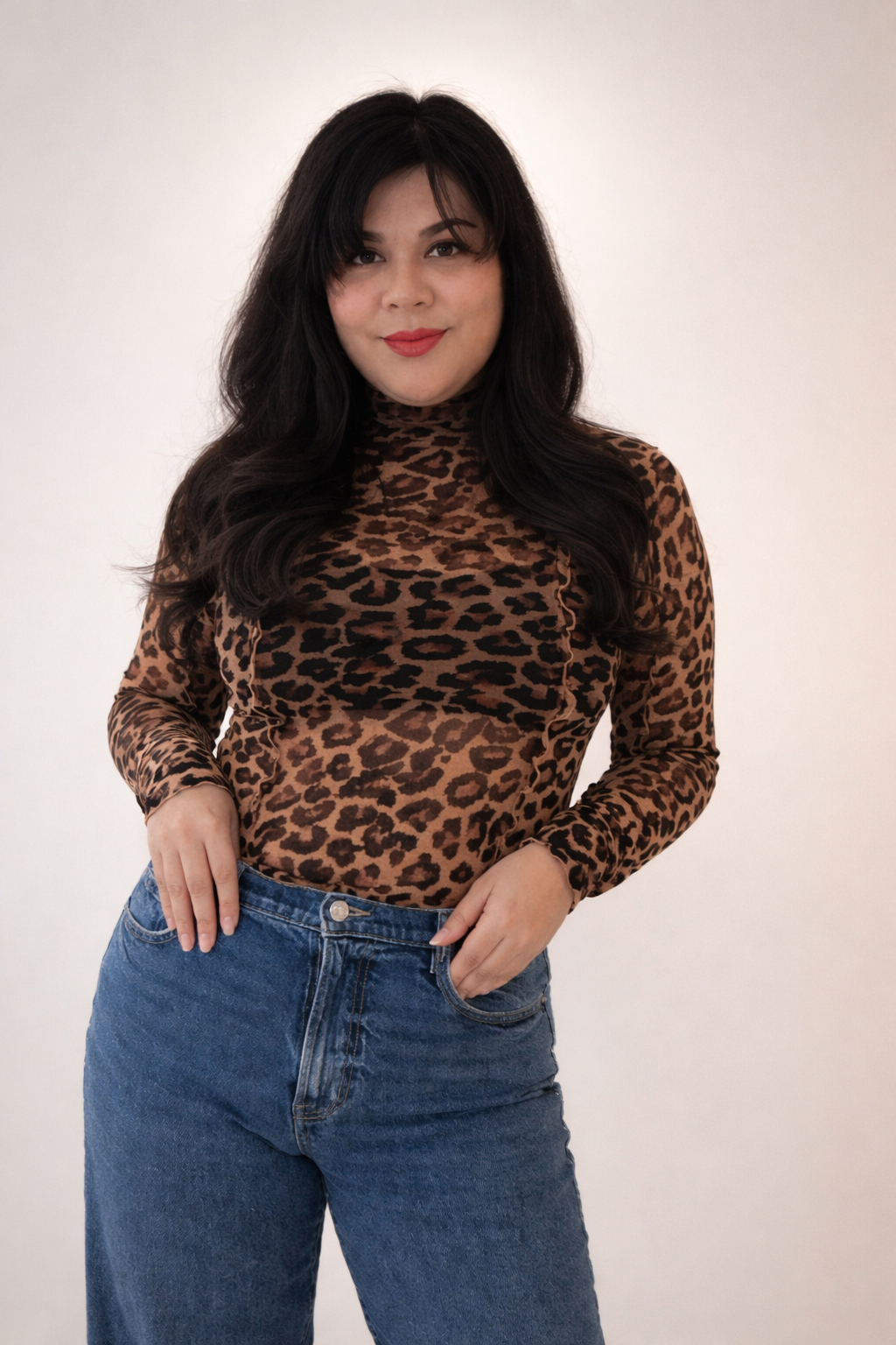 Woman with long black hair wearing a leopard print turtleneck and blue jeans, standing against a plain white background.