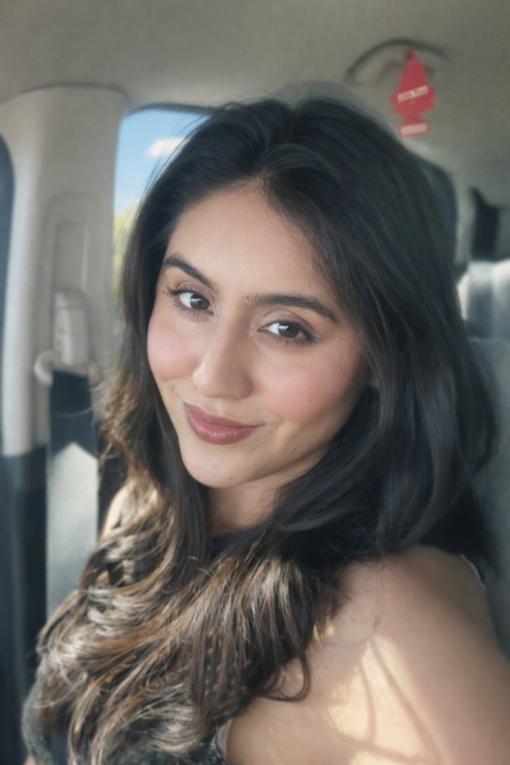 Close-up of a young woman with dark brown hair and light makeup sitting in a car, smiling at the camera.