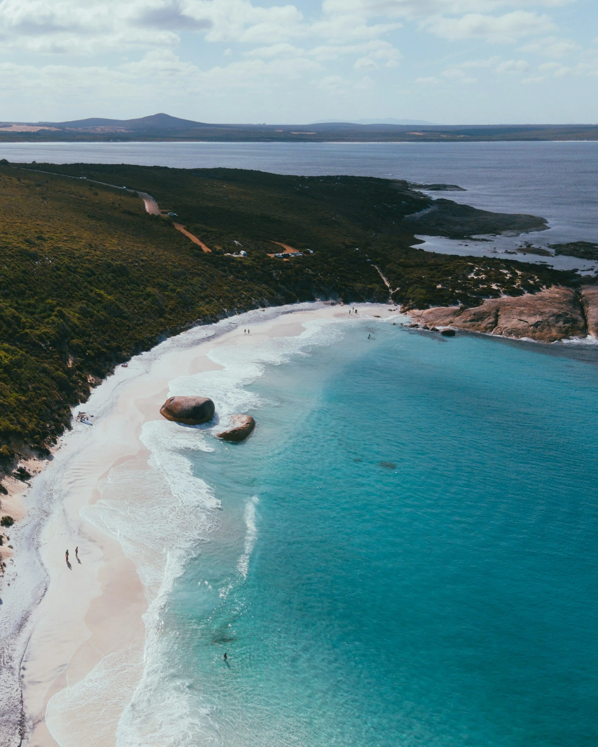 Aerial view of a beautiful beach with white sand, turquoise water, large rocks, and people swimming and walking. Green hills and a distant body of water with mountains in the background.