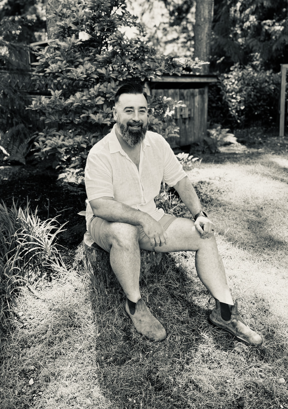A man sitting on a tree stump in a garden, smiling, wearing a light-colored shirt, shorts, and boots, with trees and bushes in the background.