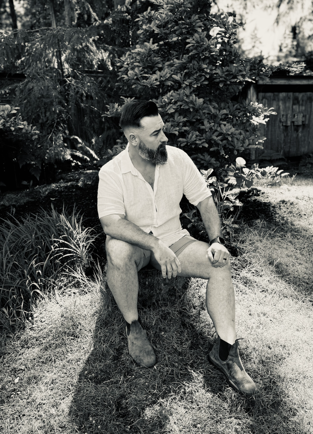 A man with a beard and slicked-back hair, wearing a light-colored shirt, shorts, and boots, sitting on a park bench in a garden with plants and trees, during daytime, in black and white.