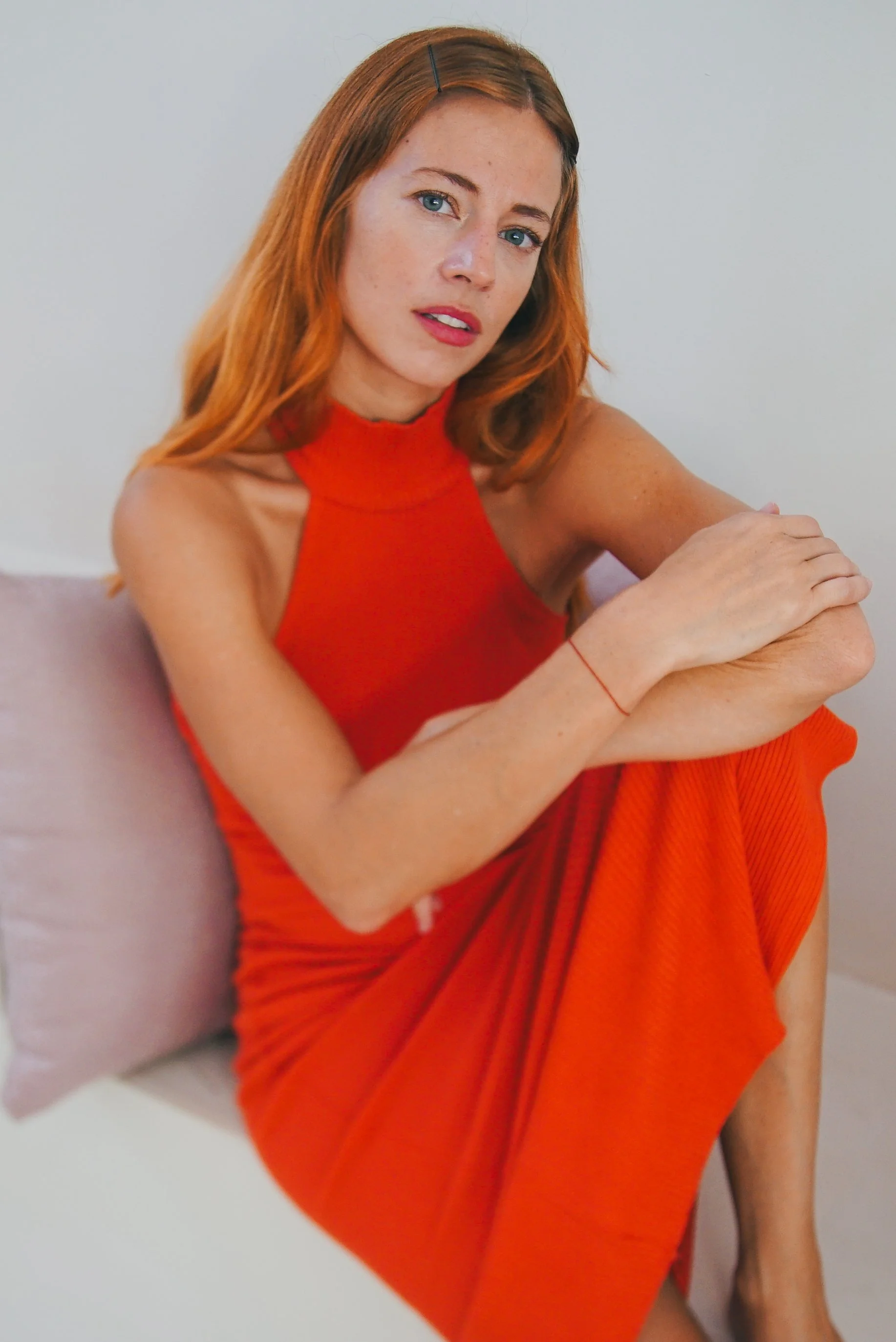 Red-haired woman in an orange dress sitting on a beige sofa, looking at the camera with a neutral expression.