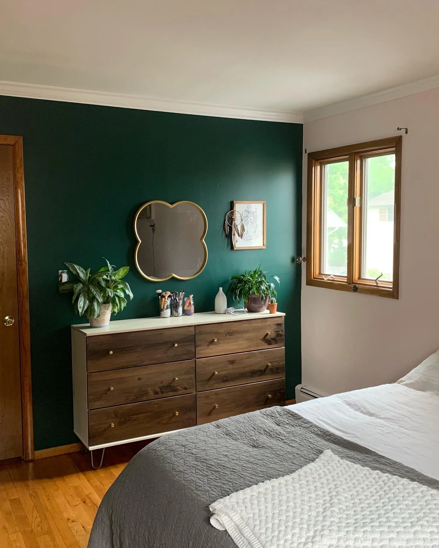 photo of a small apartment bedroom. bedroom refresh features a dark cool green wall, a gold spared mirror above the painted and stained wood dresser. light pink accent wall behind the bed.