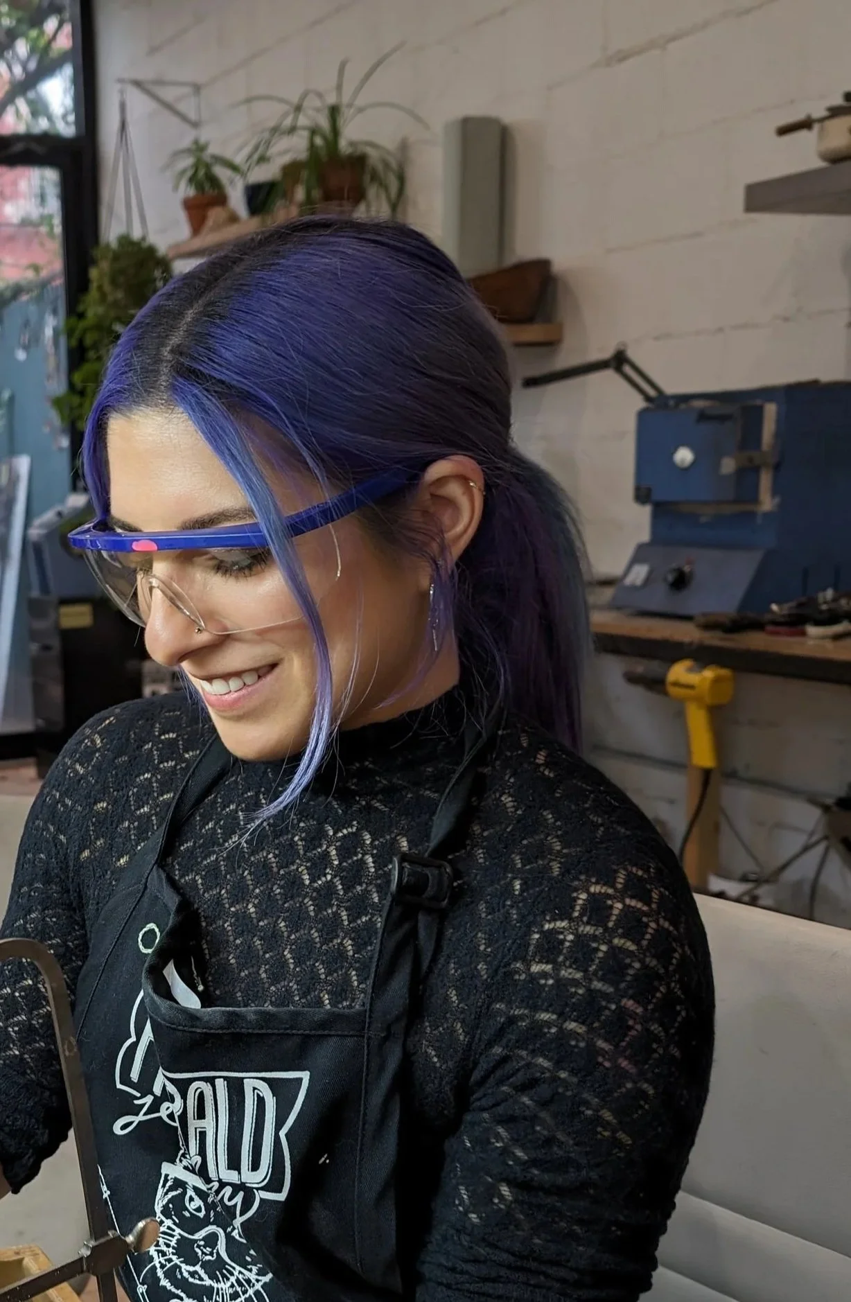 photo of myself, Kim the owner, with blue rimmed goggles, smiling and looking down. I am light-skinned woman with long dark blue hair. Wearing a long sleeve black shirt.