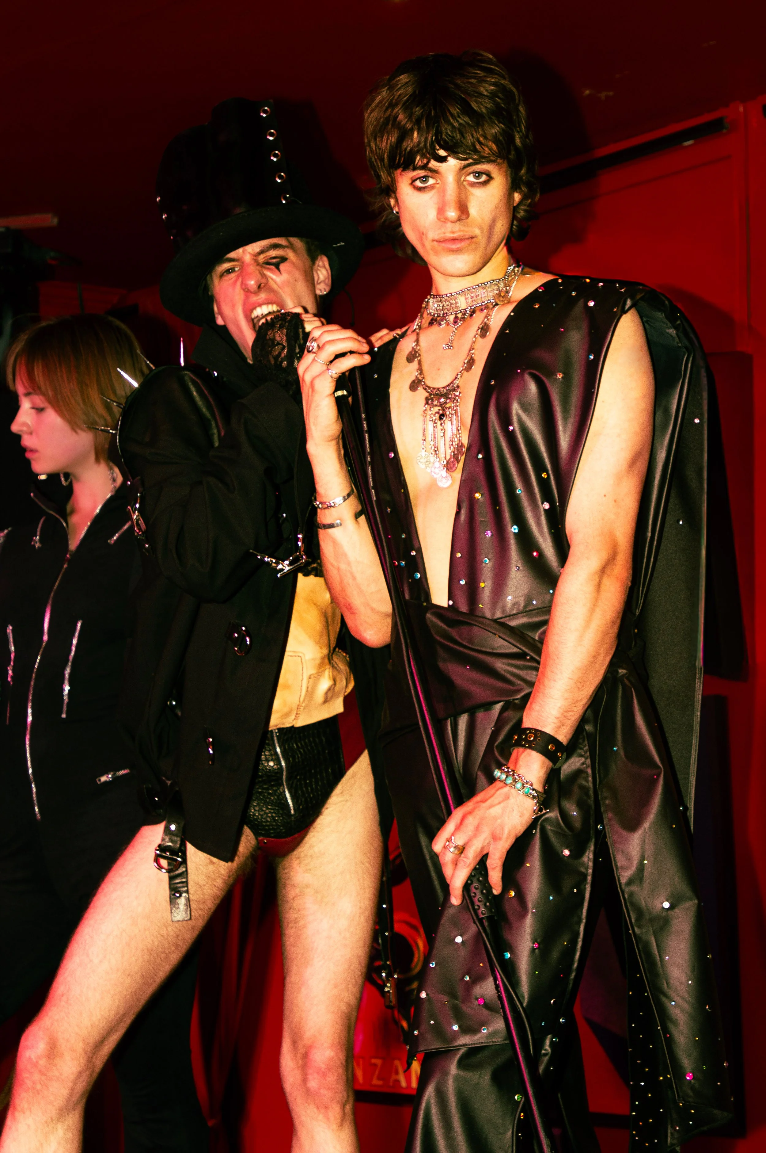 Two men in flamboyant outfits, one with makeup, jewelry, and a leather dress, in a red-lit environment with a woman in the background.