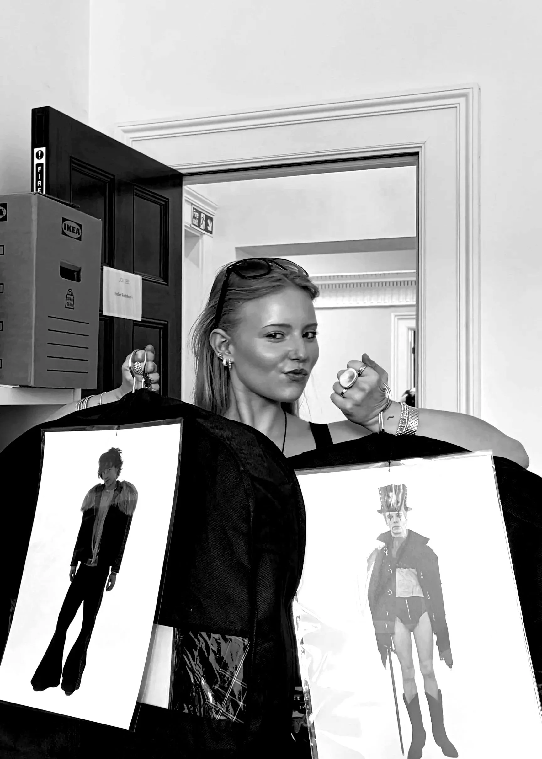 A woman standing beside black display boards featuring images of male models in edgy outfits. She is holding a set of keys and wearing jewelry, sunglasses on her head, and making a fist gesture.