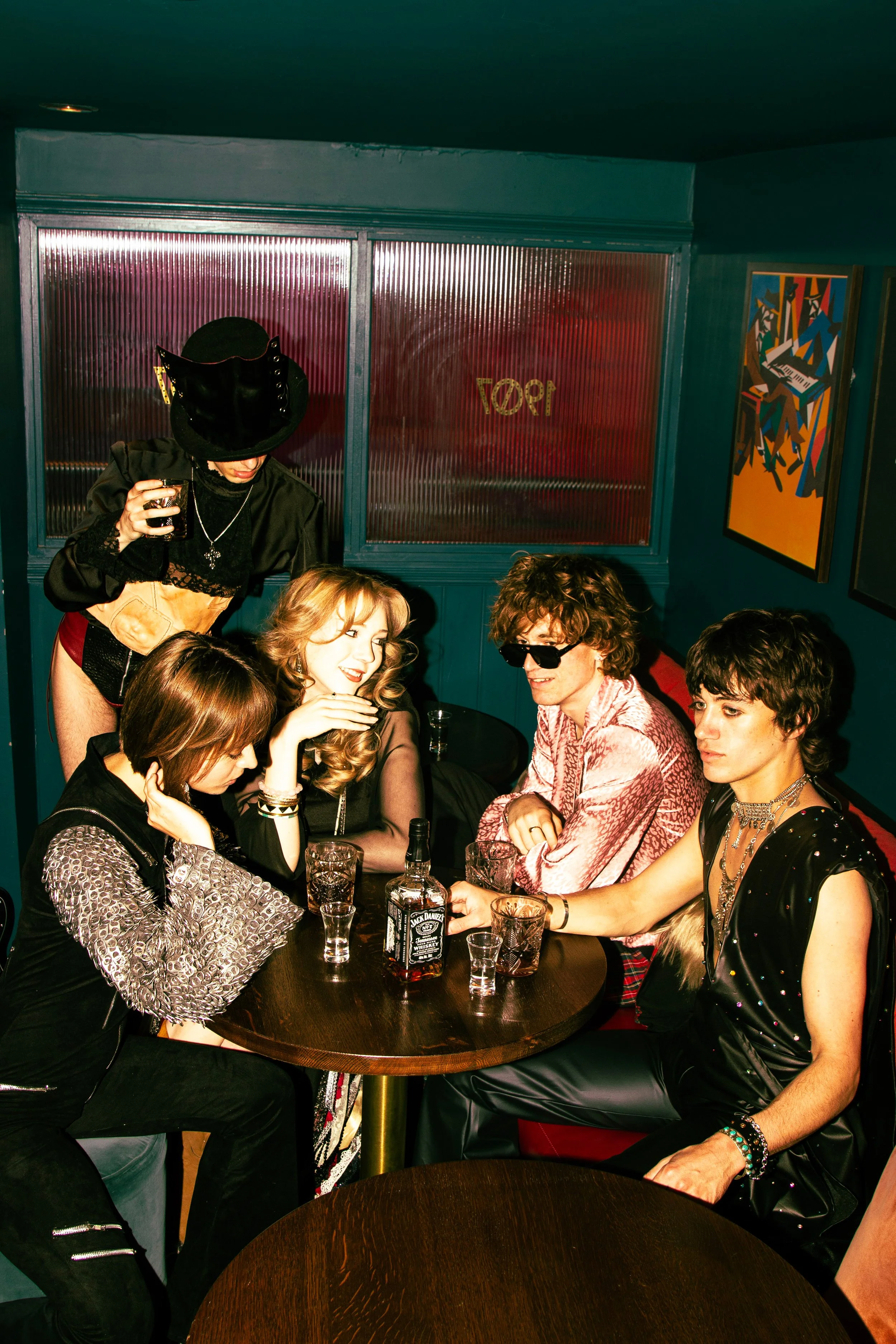 A group of six young adults sitting around a table in a dimly lit bar or club, with one woman standing, dressed in stylish and edgy clothing, drinking from a glass, with a Jack Daniel's whiskey bottle on the table. They are engaged in conversation, a
