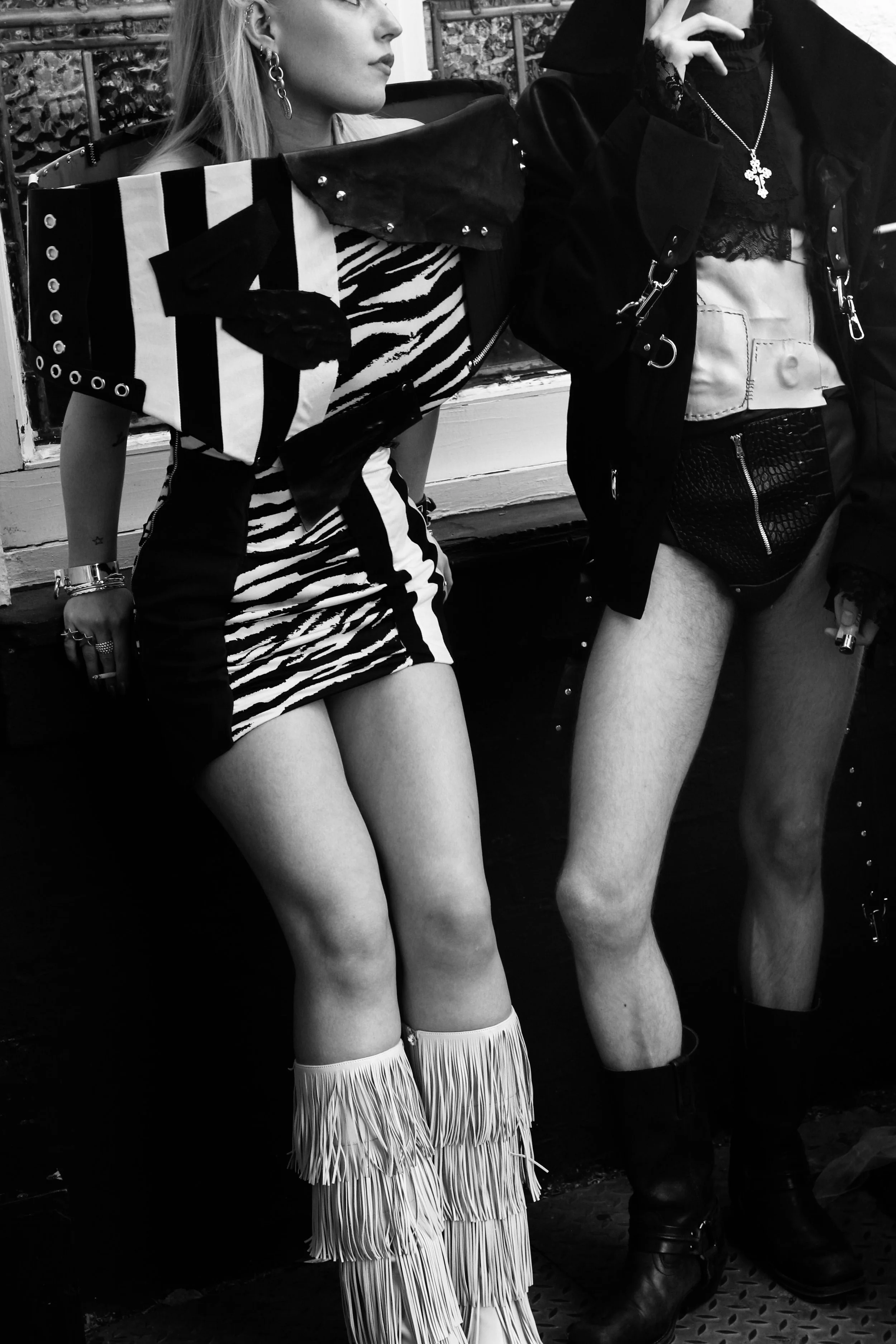 Two women sitting on a bench wearing edgy and fashionable outfits, including animal print and fringed clothing, with jewelry and accessories. One woman has long hair and is wearing a striped dress with fringed boots, while the other has shorter hair 