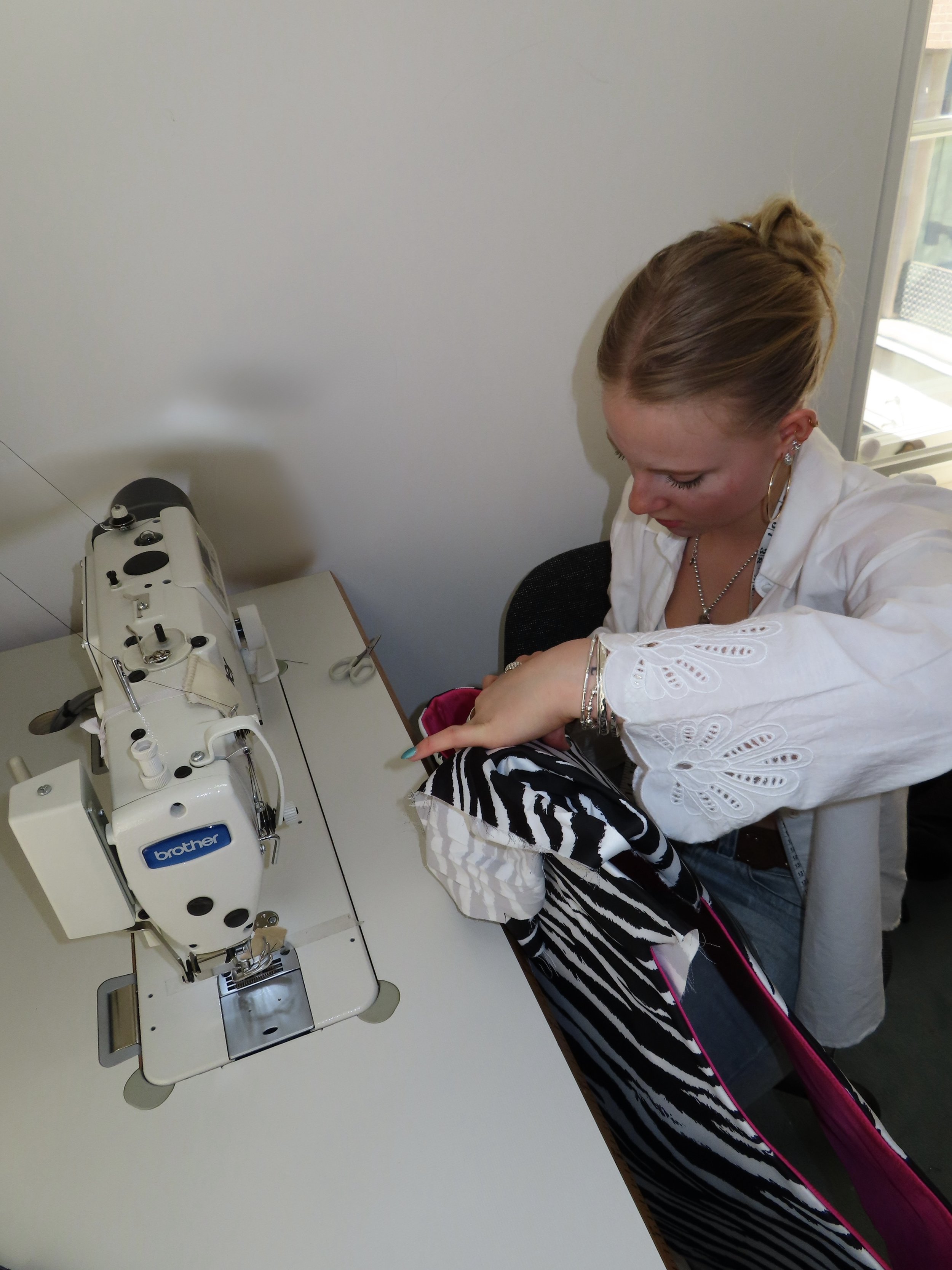 A woman sitting at a table, operating a sewing machine. The sewing machine is white with the brand name Brother visible. The woman has blonde hair tied up and is wearing a white embroidered blouse and jewelry. She appears focused on sewing a black an