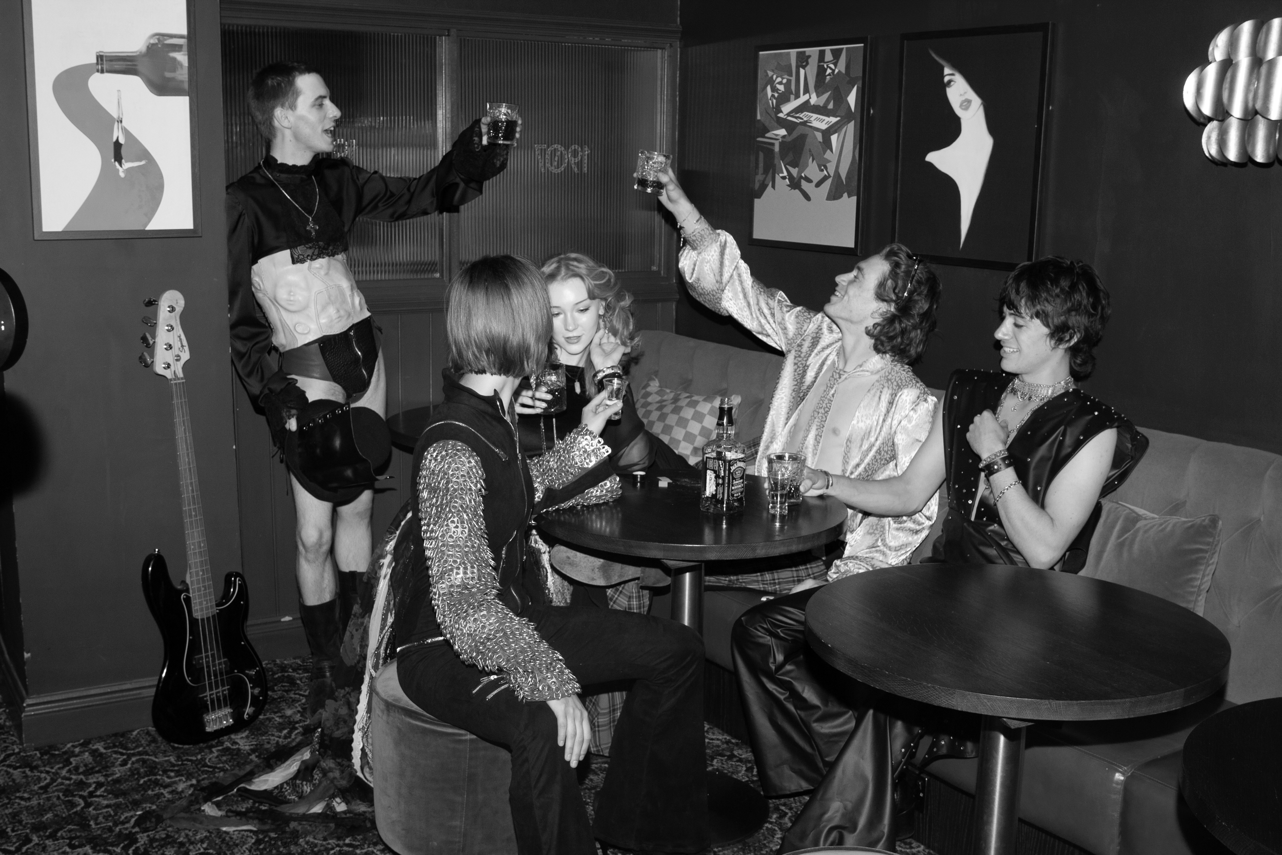A group of five young adults sitting around a table in a bar, raising glasses in a toast. The room has dark walls with modern artwork and a guitar against the wall.
