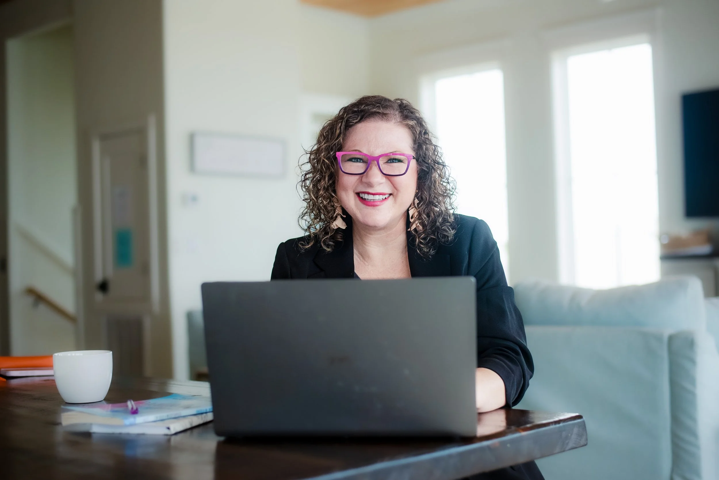 A woman with curly hair and pink glasses smiling while sitting at a table with a laptop in front of her. A white mug and some papers are on the table. The background shows a bright room with large windows.