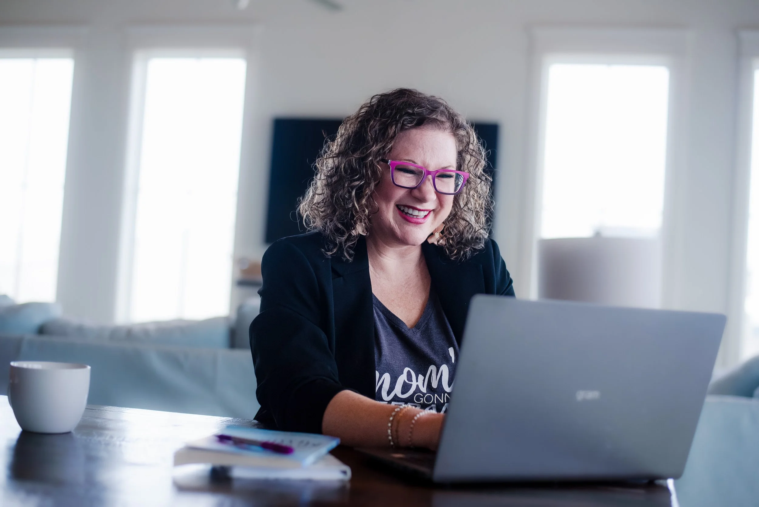 A woman with curly hair and purple glasses smiling while working on a laptop at a desk, with white coffee mugs and a notebook on the table, in a bright room with large windows.