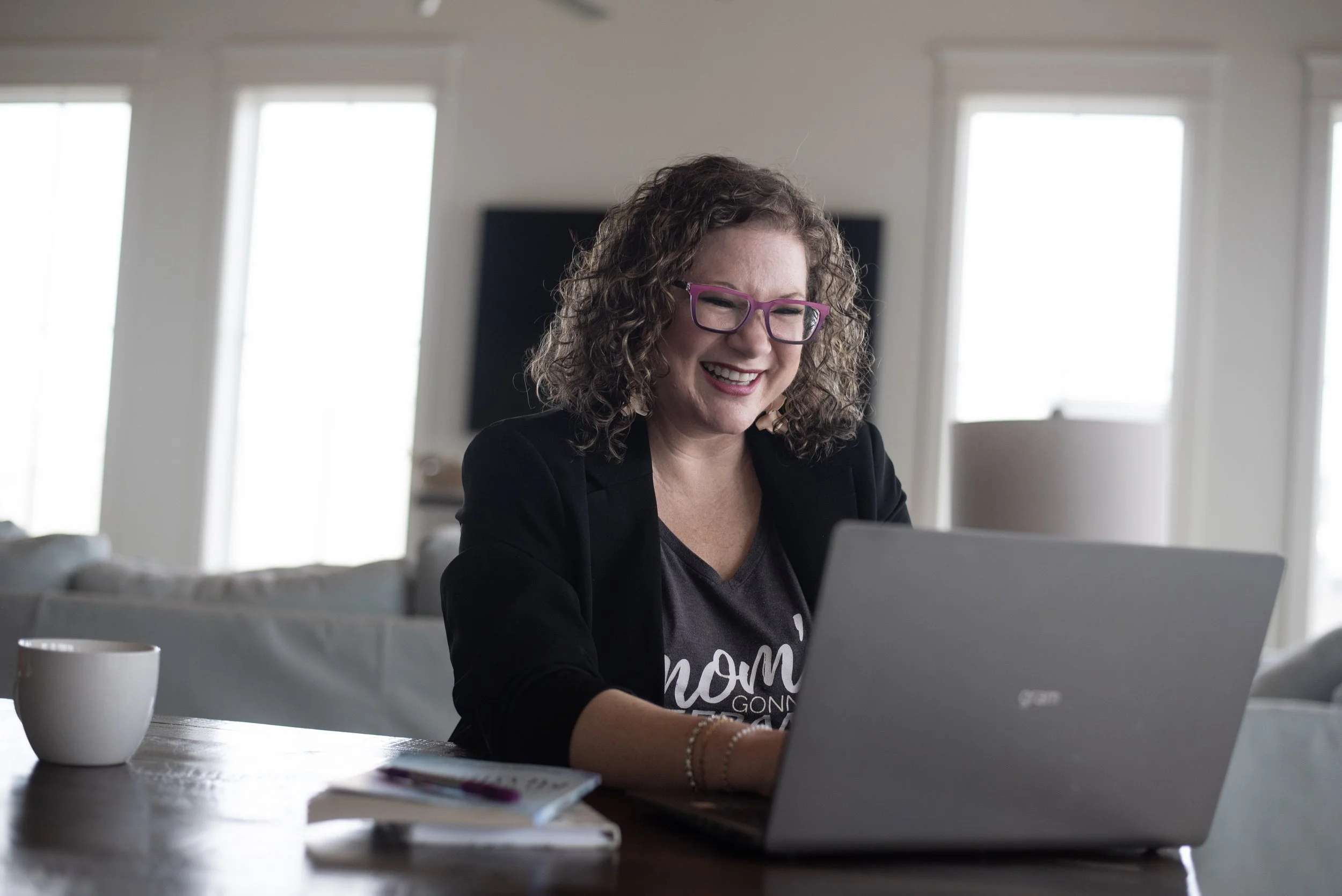 Smiling woman with curly hair and pink glasses working on a laptop at a wooden table, in a bright room with large windows.
