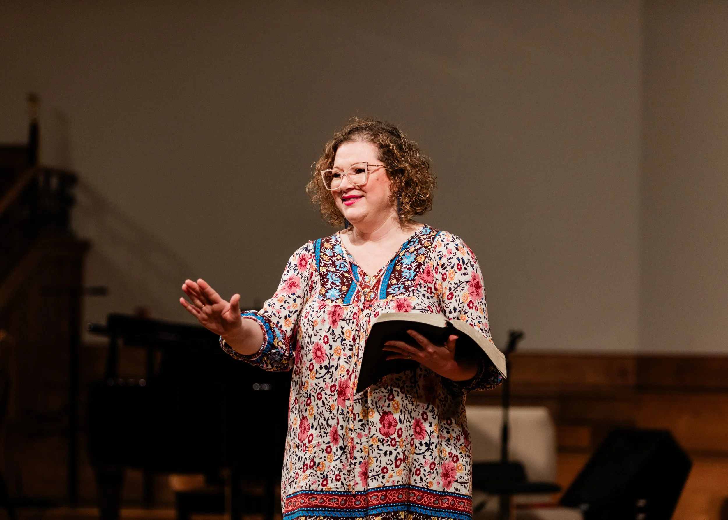 A woman with curly hair, glasses, and a colorful floral dress is holding a book and gesturing with her hand, possibly speaking or teaching in a room with musical instruments visible in the background.