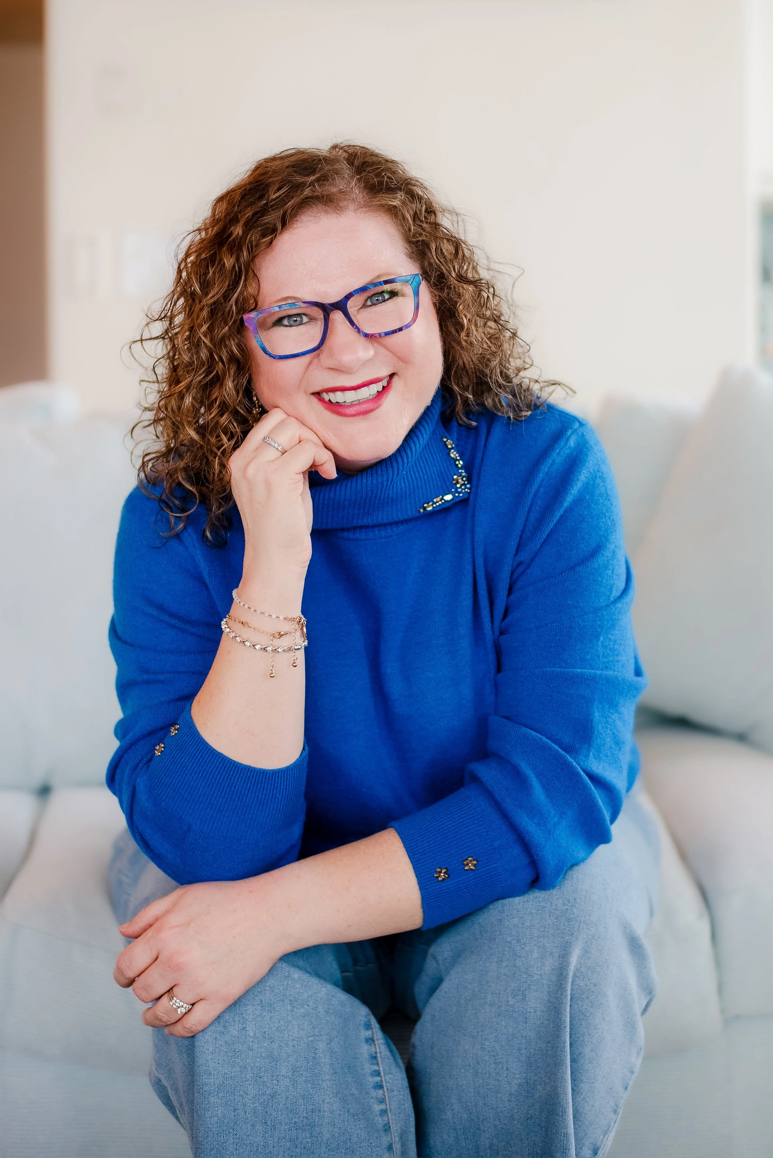 Smiling woman with curly brown hair and blue glasses wearing a bright blue sweater