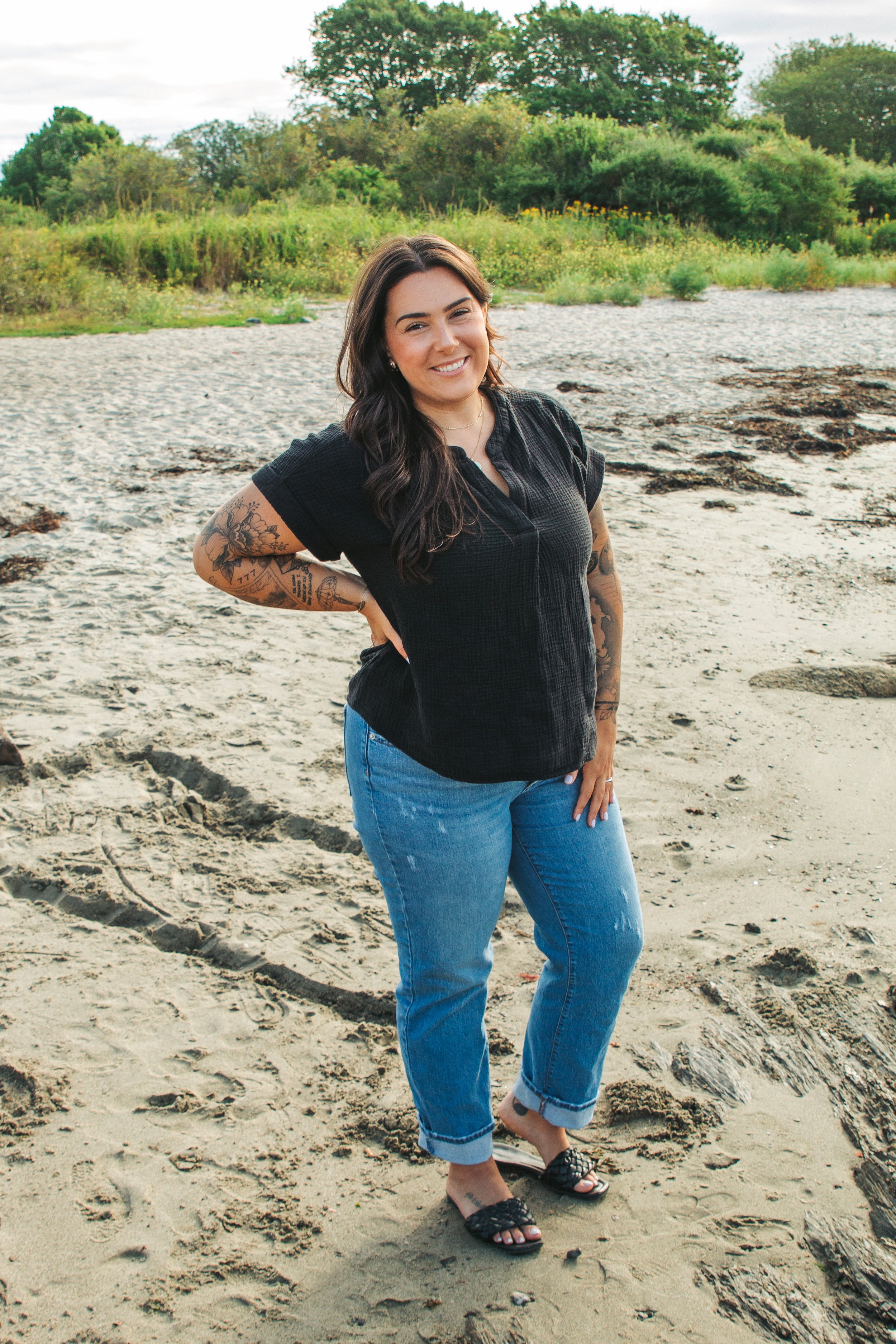 A woman with dark hair and tattoos on her arms standing on a sandy beach with greenery and trees in the background. She is smiling, wearing a black short-sleeve shirt, blue jeans, and black sandals.