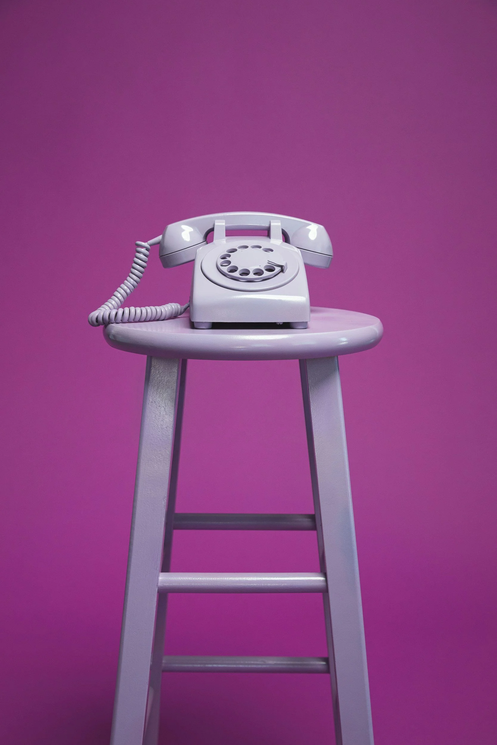 A vintage white rotary telephone on a small purple stool against a purple background.